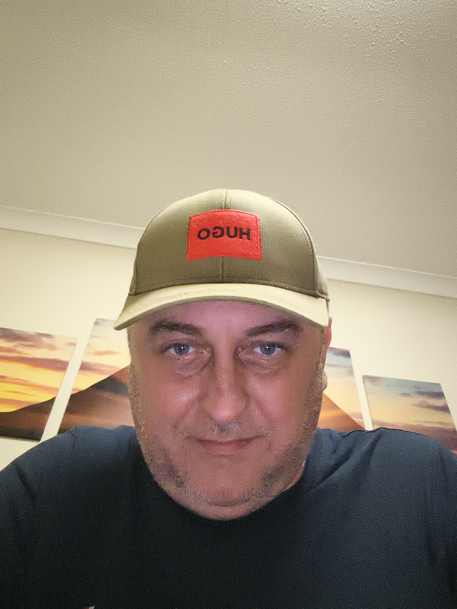A close-up, head-and-shoulders portrait captures a man indoors, likely a selfie. He appears to be middle-aged, with blue eyes and short facial hair/stubble, sporting a neutral to slightly smiling expression. He wears a dark, possibly navy or black, t-shirt. On his head is an olive green baseball cap featuring a distinctive rectangular red patch on the front. White capital letters on this patch read "HUGO," but are inverted in the image, appearing as "OƆUH."
The background reveals a light-colored wall and ceiling, with a visible molding strip at their junction. Behind the man, framed art prints or canvases depicting warm, possibly sunset or sunrise landscapes, add color to the domestic setting. The lighting is artificial, emanating from above and providing an even illumination. The man is simply posing for the camera. Based on the provided context, the scene is located in Huntingdonshire, United Kingdom. The time of day is not discernible from the indoor lighting.