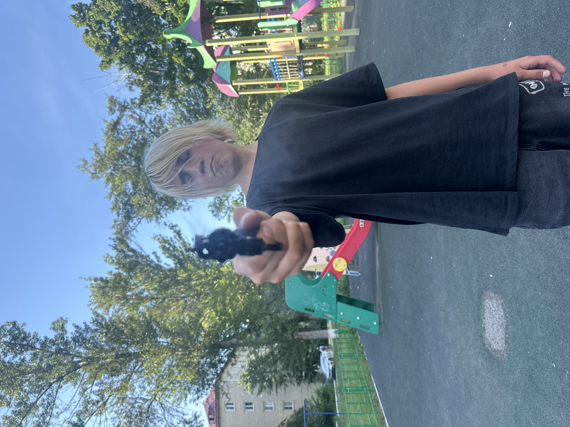 A young person with bleached blonde hair, wearing a black t-shirt and dark pants, stands in what appears to be a city playground in Togliatti, Russia. They are holding a dark-colored handgun in their right hand, pointing it directly at the camera. The playground features brightly colored slides and climbing structures, along with a paved surface and a green fence. Trees and a multi-story building are visible in the background, under a clear blue sky. The overall impression is one of an intentionally staged photograph.
