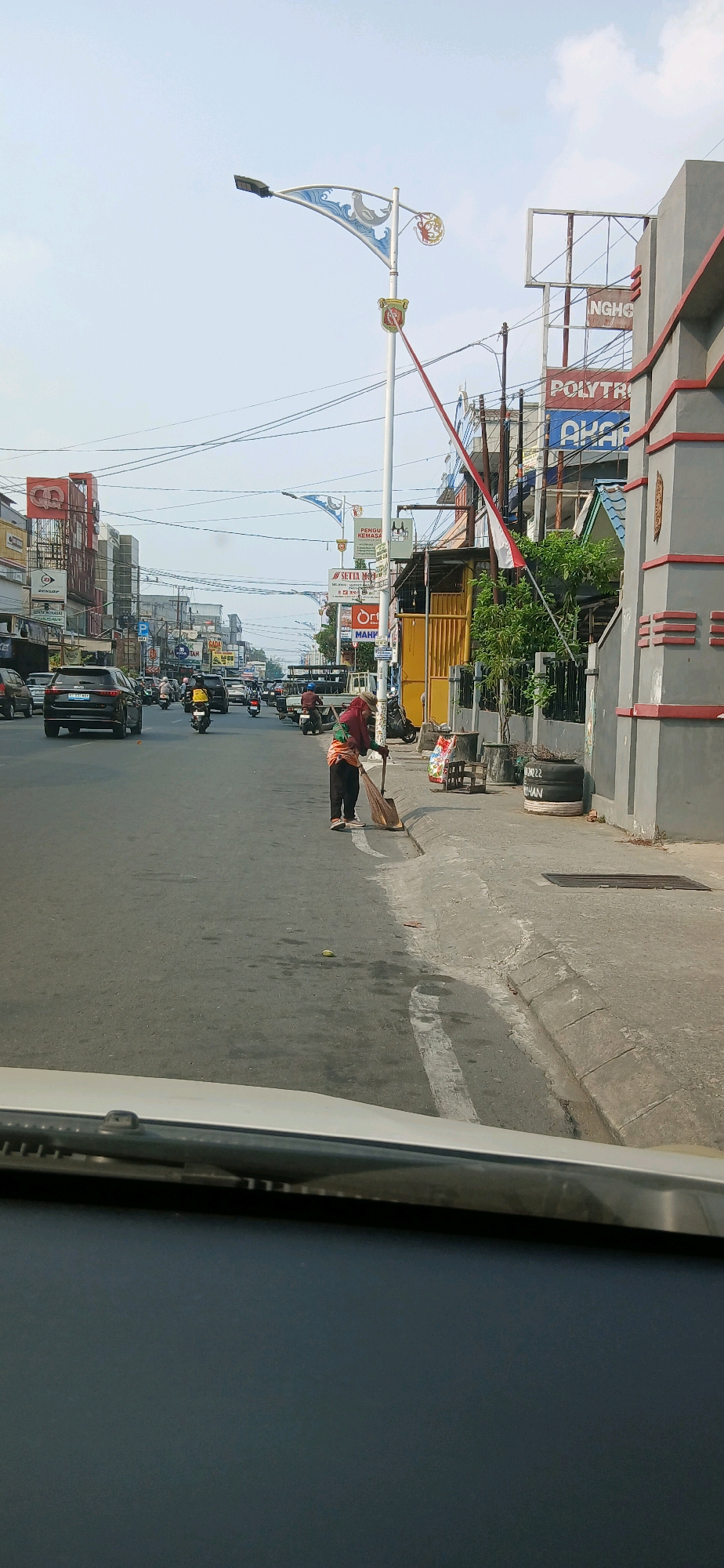 In Samarinda, Indonesia, a person is sweeping the sidewalk next to a street with moderate traffic. The person, wearing a head covering, long-sleeved shirt, and dark pants, uses a broom to gather debris. The street has several cars and motorcycles. Buildings line the street, featuring advertisements and signs in Indonesian. A white car's dashboard and windshield are visible in the foreground, indicating the photo was taken from inside a vehicle. The sky is clear and bright.