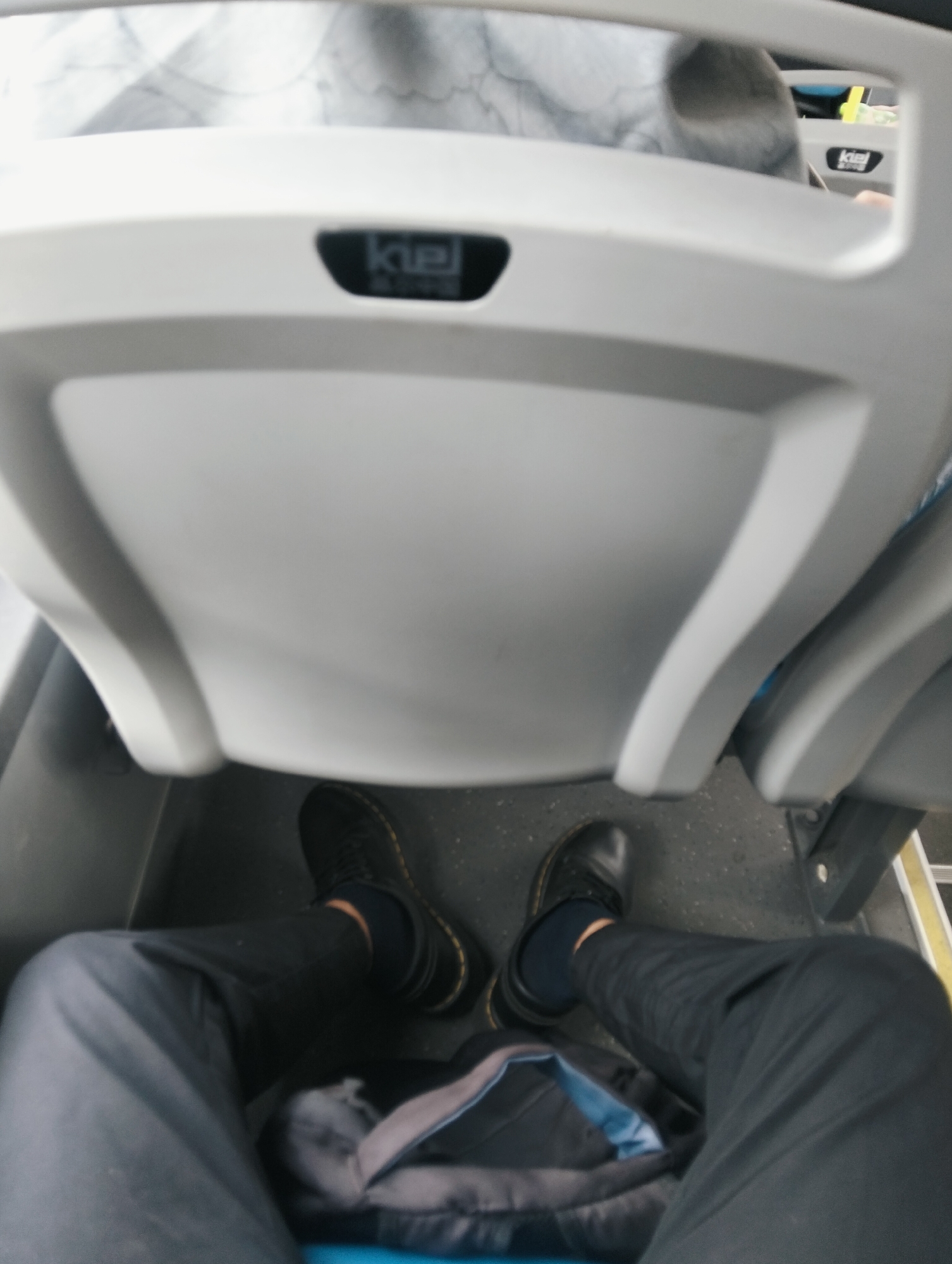 The image is a first-person perspective shot looking down at the viewer's legs and feet while seated on a bus. The viewer is wearing dark pants and black lace-up boots with yellow stitching. A backpack is partially visible between the viewer's legs. The bus seats are white and grey, and a logo with the word "kiel" is visible on one of the seats. The floor of the bus is grey and speckled. The image appears to have been taken in a public transportation setting. There are no identifiable landmarks or contextual clues that suggest the location is specifically the City of Medan, Indonesia.