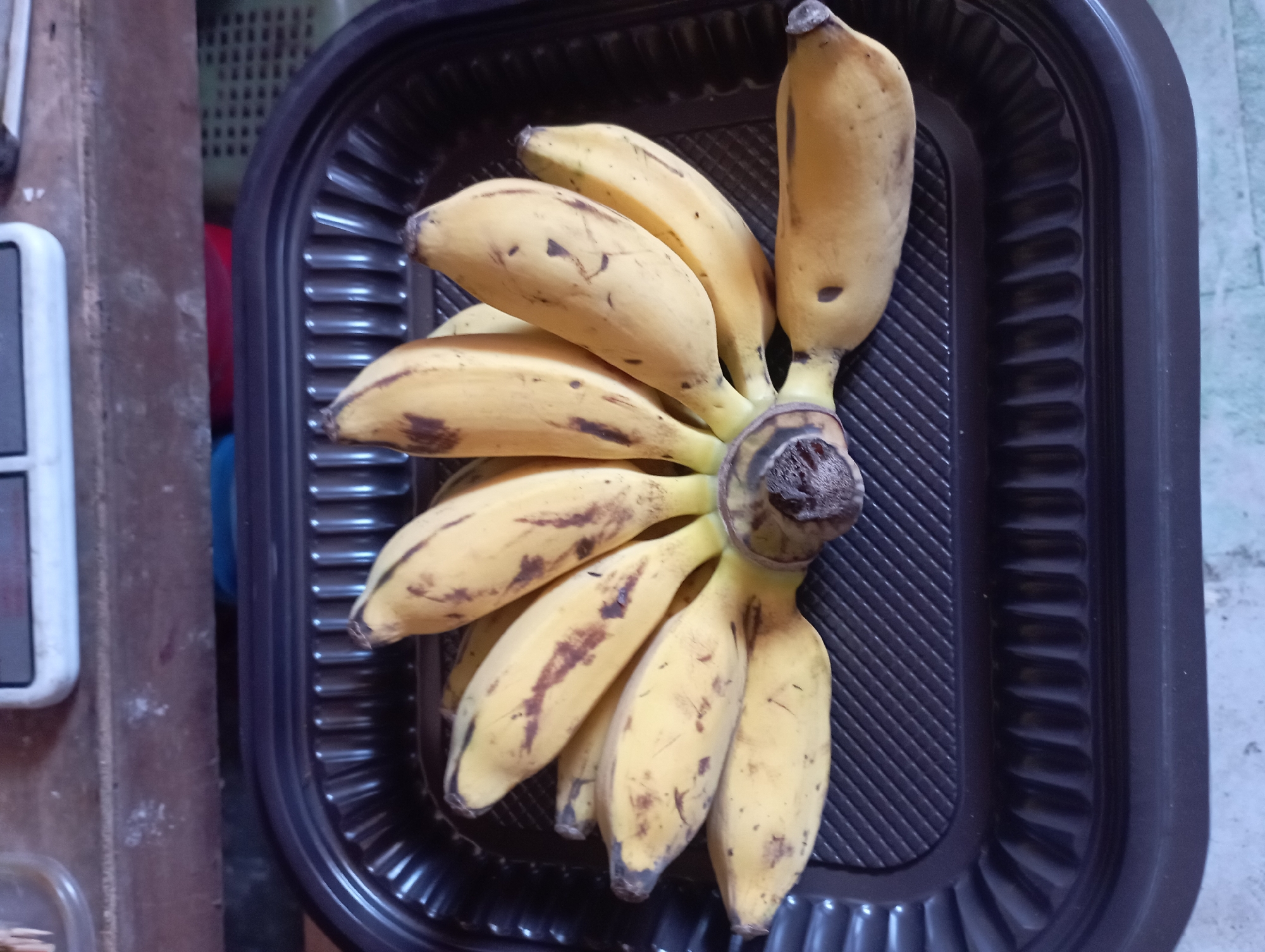 In Sembung Anyar, Indonesia, a bunch of yellow bananas with brown spots is arranged in a black plastic tray. The tray has a ridged pattern on its surface. The bananas are clustered together, with some slightly overlapping. The background shows a wooden surface and a white electronic device. The lighting appears to be natural, casting soft shadows.