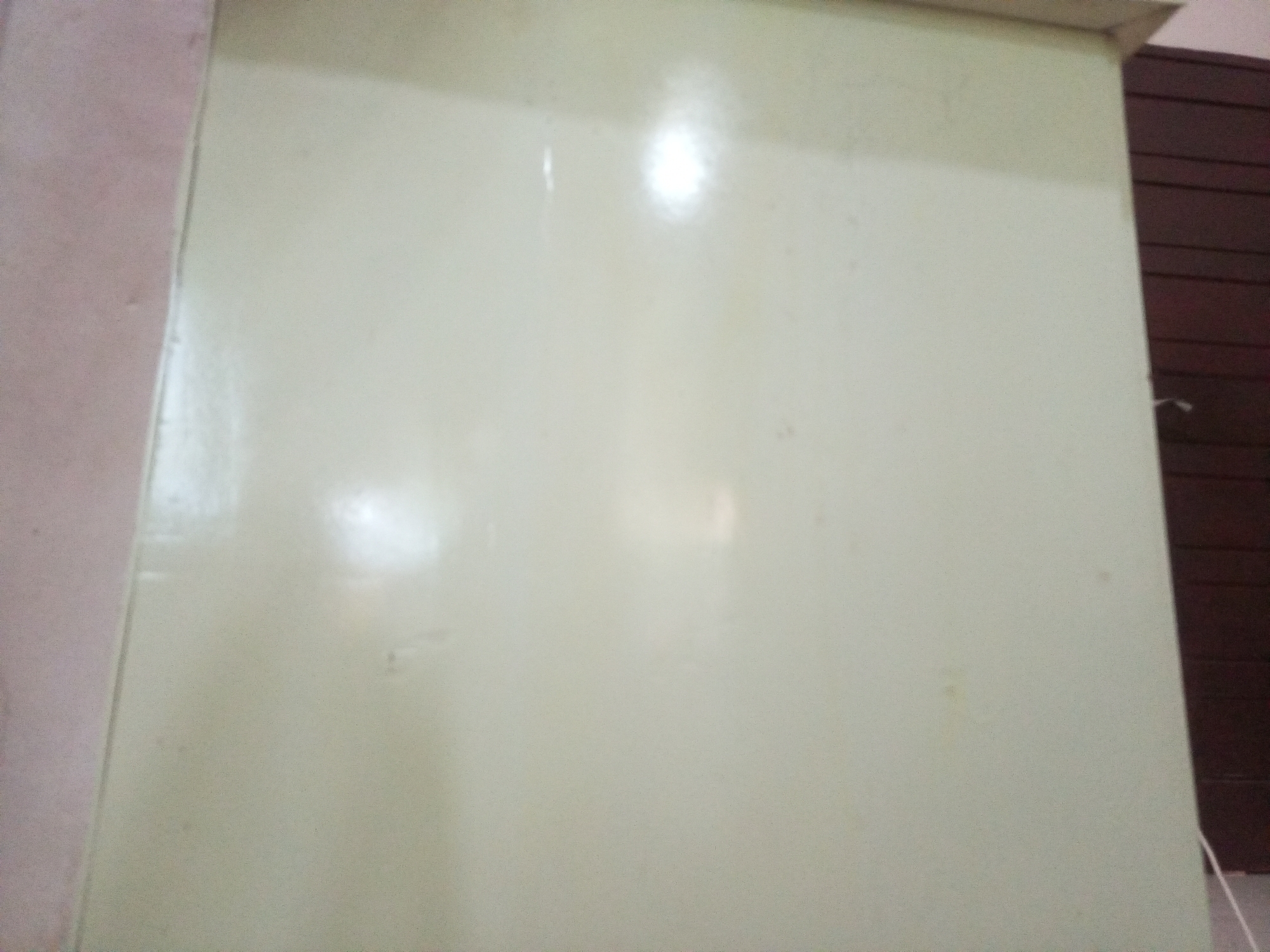 The image shows the side of a light-colored cabinet or refrigerator, likely in Lhokseumawe, Indonesia. The cabinet has a smooth, slightly reflective surface, with some glare from overhead lighting. To the left of the cabinet, a pinkish wall is visible. To the right, a dark brown wooden surface with horizontal slats is present, suggesting it could be a door or furniture. A thin white cord or wire is faintly visible running along the bottom right edge of the frame. There are no people or clear actions depicted in the scene. The environment appears to be indoors.