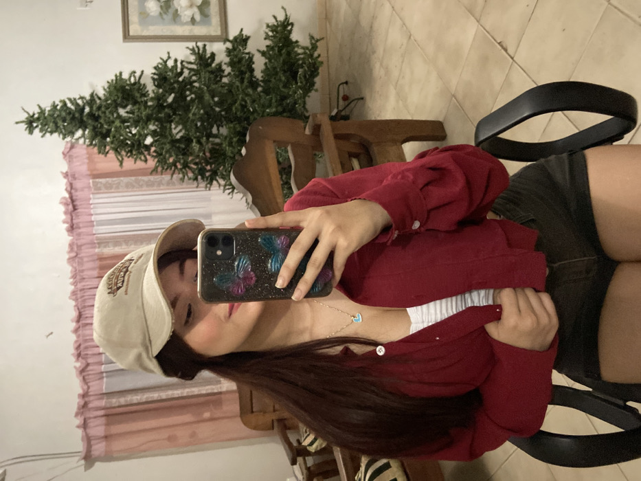 A young woman poses for a selfie. She wears a tan baseball cap, a white top under an open red collared shirt, and dark shorts. She holds a smartphone with a butterfly-patterned case. She sits in a black office chair. Behind her, a small artificial Christmas tree stands near a wooden chair, in front of a window with light pink curtains. The room has light-colored tiled floors, and there is a framed painting on the wall. The image is rotated clockwise.