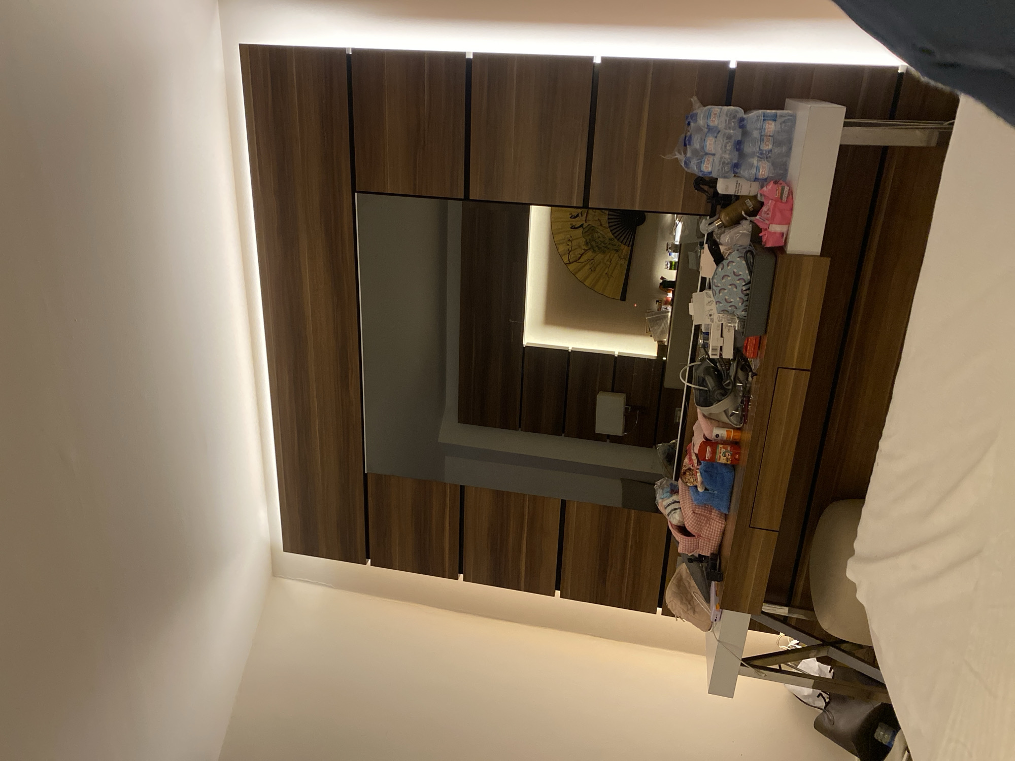The image depicts a dimly lit hotel room, possibly in Aksu, Turkey, with a focus on a dressing table and a large wooden headboard with an integrated mirror. The dressing table surface is cluttered with various items including toiletries, a pink makeup bag, a stack of bottled water, and other personal belongings. A partially visible beige pouf is situated in front of the table. The headboard features a large mirror reflecting a yellow fan with avian imagery and some ambient lighting from within the unit. The overall environment suggests a domestic setting, likely a hotel room preparing for occupancy or having been recently used. The lighting is soft and ambient, with vertical strip lights flanking the wooden headboard. There are no people clearly visible in the frame.