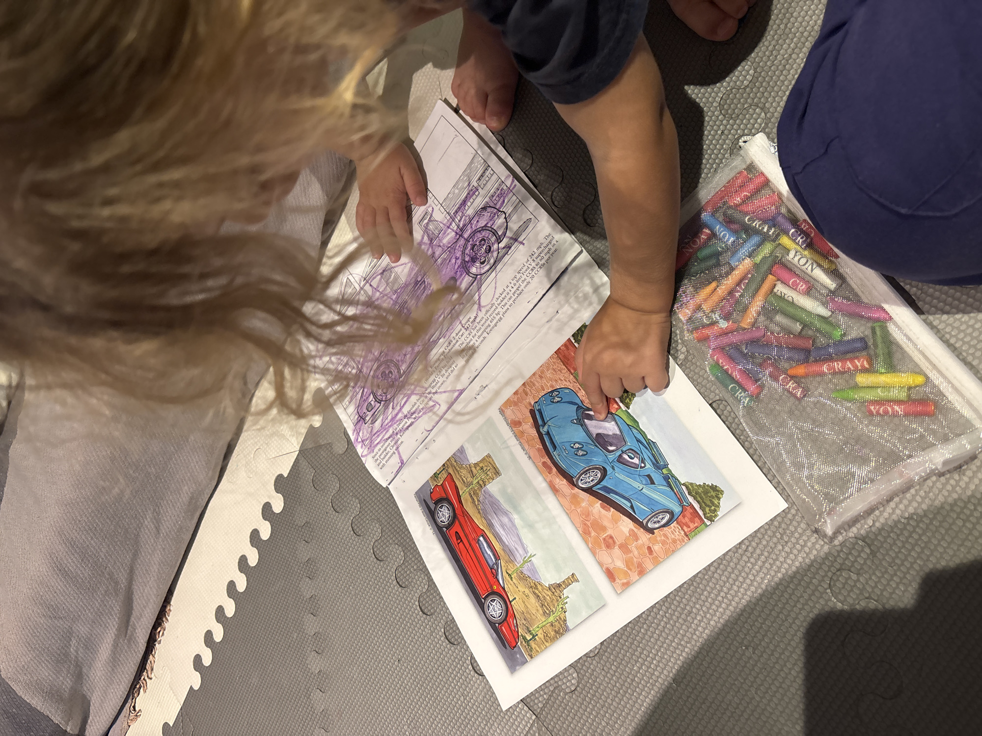 An overhead shot shows a child with blonde hair, sitting on a grey mat with puzzle-piece-like indentations. The child is looking down at an open book with colorful illustrations of cars. One page has a red car on a rocky cliff, and the other has a blue car on a brick road. Next to the book, a clear plastic pouch is filled with various colored crayons. The child's hands are visible, one resting on the blue car illustration, and the other holding a crayon. The child is wearing a dark blue shirt. The background features a folded grey blanket and a portion of a light-colored mat. The scene appears to be indoors, likely a home setting, and there is no indication of a specific city or location like Dubai.