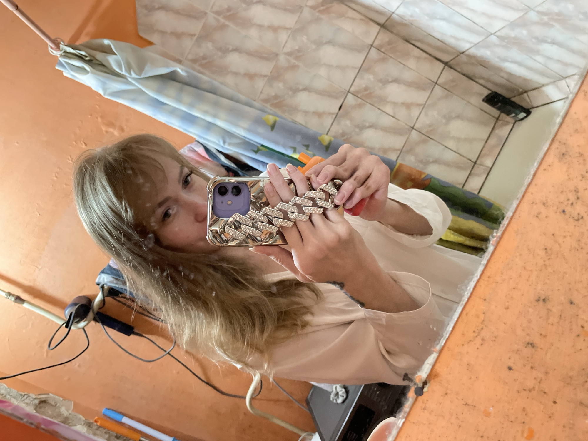 A person is taking a selfie in a bathroom mirror. The phone case is decorated with a diamond-like pattern. The bathroom has orange walls and tiled shower walls. The location is not discernible from this image.