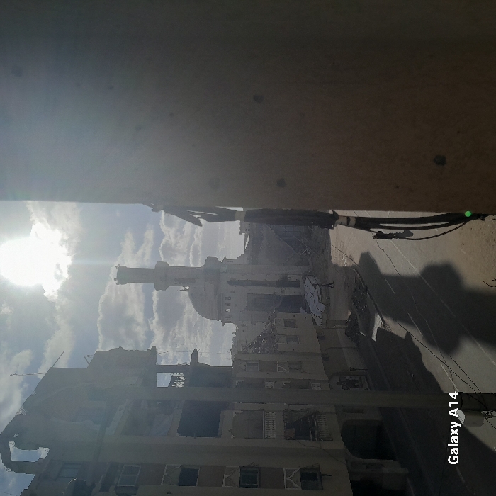 The image depicts a war-torn city landscape under a bright sun. Buildings are heavily damaged with gaping holes and rubble visible. A mosque with a prominent minaret and dome stands amidst the destruction. Shadows stretch across the street. The photograph seems to be taken from a higher vantage point, perhaps a building overlooking the scene. "Galaxy A14" is printed on the bottom right corner, indicating the device used to capture the image.