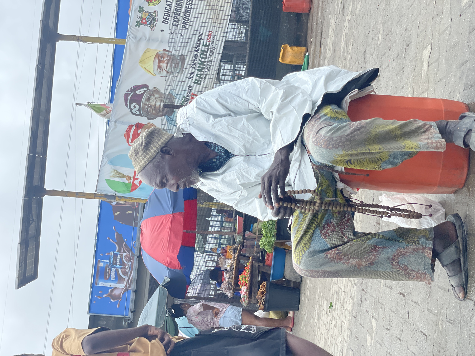 In Ajah, Nigeria, an elderly Black man with a beard and a patterned knit cap sits on an orange plastic drum in an outdoor market. He wears a white robe and vibrantly patterned trousers. His hands are clasped together, holding a string of prayer beads. Behind him, a banner features portraits of three men in traditional caps, with text that appears to be political campaign material. Various market stalls are visible, including one with produce like peppers and leafy greens, and another with items on display under red and blue umbrellas. Other people are present in the background, including a woman in denim shorts and a patterned top. The ground is paved with interlocking bricks, and the sky is overcast.