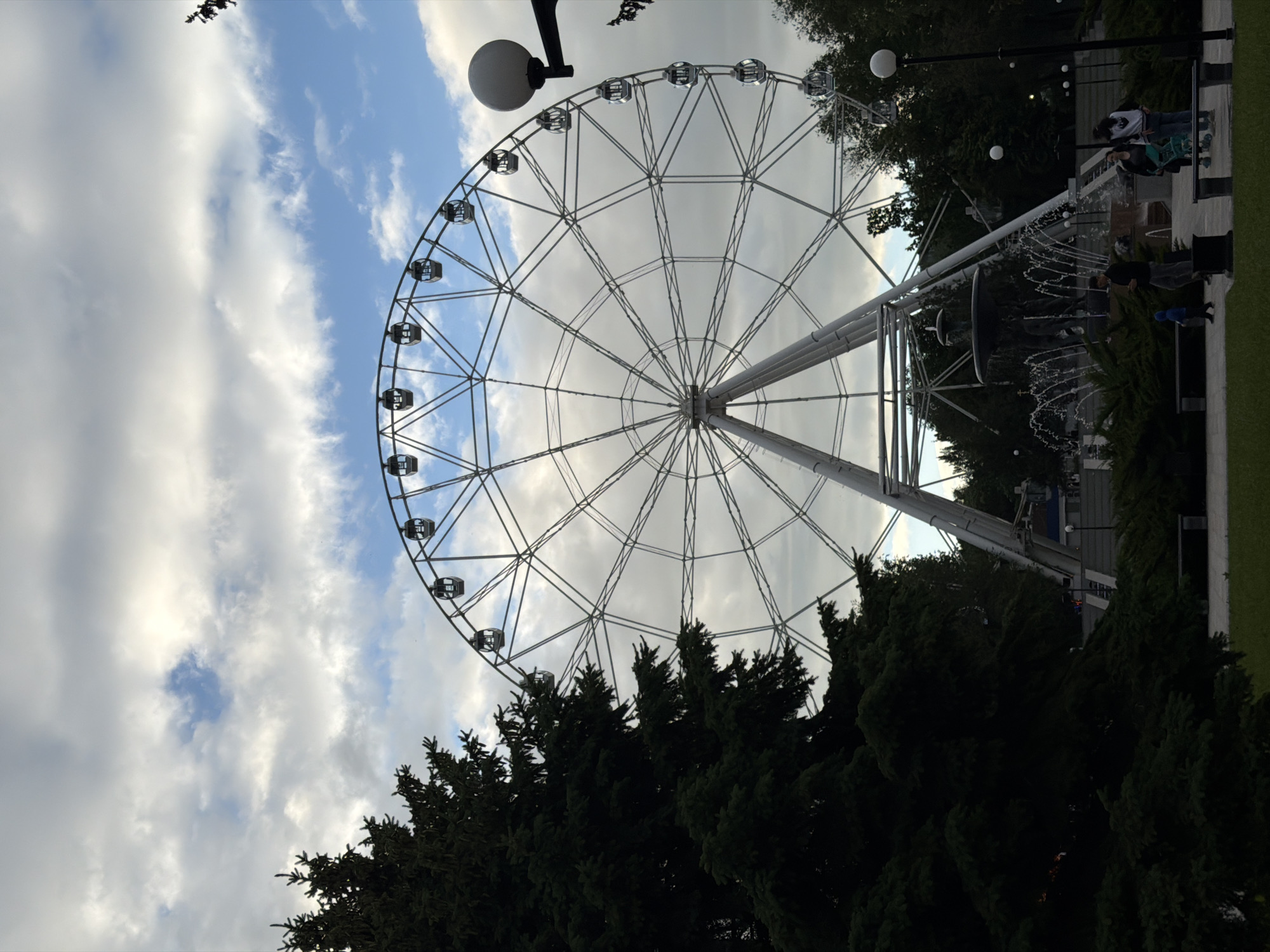 The image shows a large Ferris wheel located in Rostov-on-Don, Russia, as indicated by the city's skyline. The wheel, constructed of a white metal frame, dominates the scene, featuring numerous passenger cabins evenly spaced around its circumference. The sky is partially cloudy, with patches of blue visible. Below the Ferris wheel, there are trees and some landscaping, including bushes and possibly a fountain, which are typical features of a park or amusement area. People are visible in the background, suggesting an active public space. The image appears to have been taken from a low angle, emphasizing the size and scale of the Ferris wheel. Street lamps with spherical lights are present near the Ferris wheel.