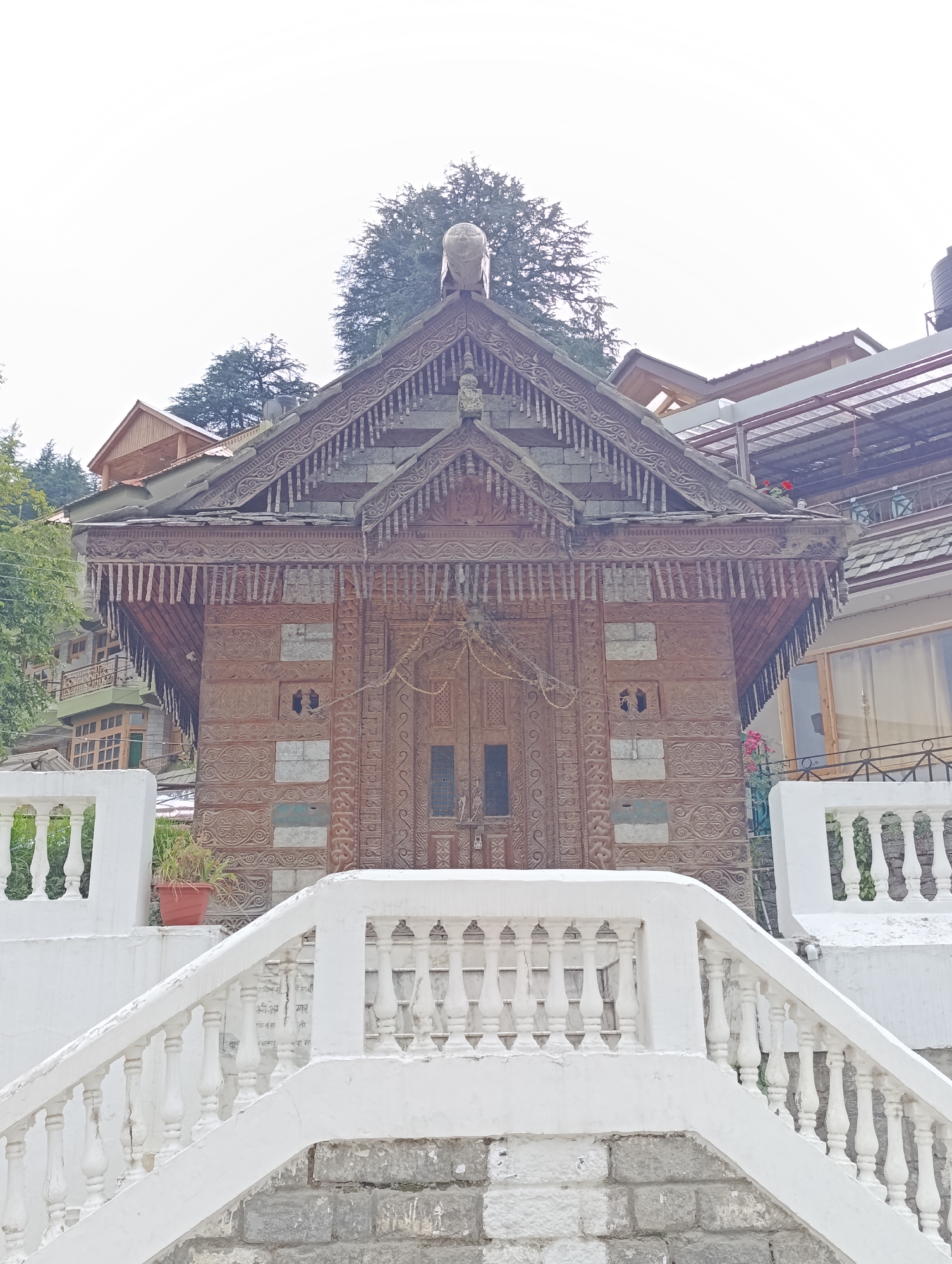 An intricately carved wooden temple or shrine stands centrally in the image, showcasing traditional Himalayan architecture. Its prominent gabled roof is adorned with slate or stone tiles and features ornate wooden eaves with decorative pendants. A round, carved finial crowns the apex of the roof. The temple's walls are constructed from alternating horizontal dark wooden beams and lighter, rectangular stone or plaster blocks. The wooden sections are covered in elaborate carvings, displaying geometric patterns, swirling motifs, and floral designs. In the center, two double wooden doors are also richly carved. Smaller, square openings or windows with intricate grillwork are visible above and to the sides of the main doors.
In the foreground, a white concrete balustrade with classic baluster shapes forms a staircase leading up towards the temple. The base of this structure is made of rough grey stone blocks. A single potted plant sits on a white ledge to the left. Faint text in Devanagari script is visible on the right-side balustrade, including phrases like "श्री आपर" and "नगा रा," likely indicating a name or dedication pertinent to the temple or location (Naggar, India).
In the background, other buildings with gabled roofs are visible behind the temple, some featuring balconies and glass windows. Lush green conifer trees dot the hillsides behind these structures, suggesting a mountainous or hilly environment. The sky is bright and overcast, indicating a cloudy day with soft, diffuse lighting and no harsh shadows. A black water tank can be seen on a distant rooftop to the right. No human activity is depicted in the scene.