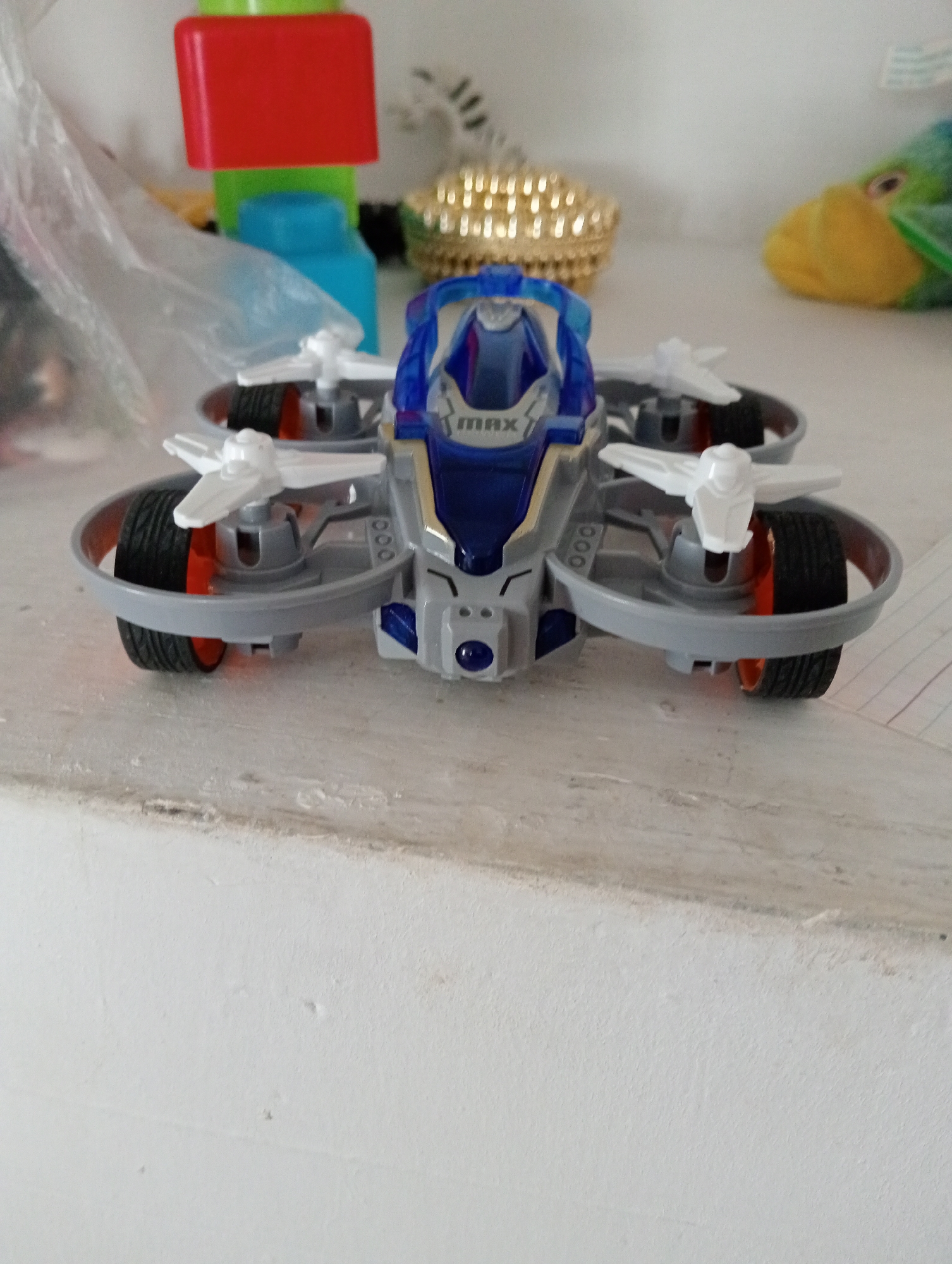 An overhead drone toy with blue and silver detailing sits on a white, textured surface. The drone has four rotors, two of which are visible in the foreground, each with a black tire and an orange inner rim. The body of the drone is sleek and futuristic, with "MAX" printed in white on the top. In the background, out of focus, are several other objects, including a red block on top of a green and blue structure, a gold, textured sphere, and a colorful plush toy of a bird with a yellow face and green body. The overall scene appears to be an indoor setting, likely a shelf or table, with the drone being the primary subject.