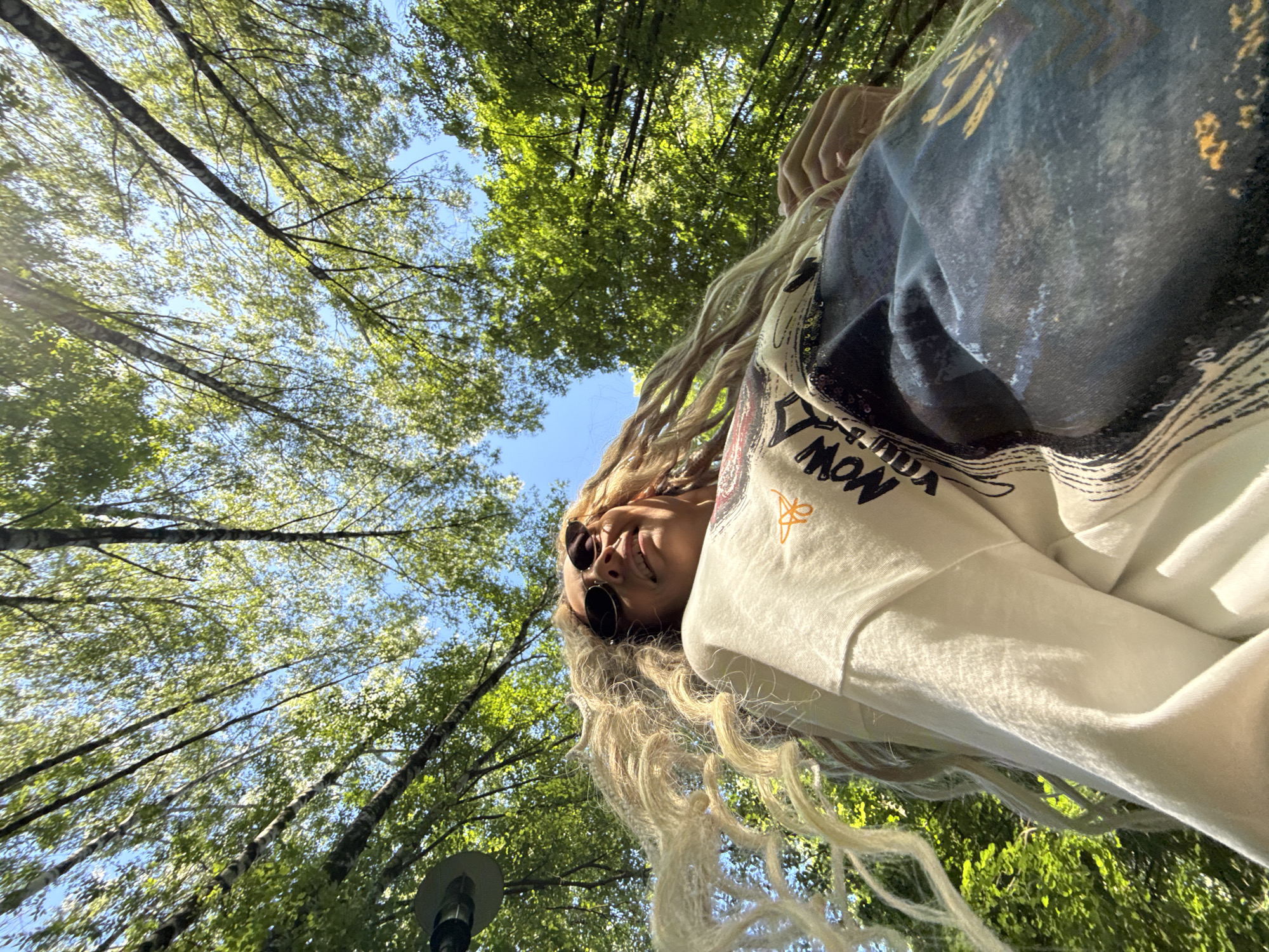 An overhead shot captures a woman with long, light-colored dreadlocks and wavy hair, wearing dark sunglasses, a white long-sleeved shirt with an orange graphic, and a dark blue or gray vest or shawl over it. Her head is tilted slightly to the right, and she is smiling. The bright blue sky is visible behind her, with green tree canopy on either side and above her, suggesting a forest or park setting. Sunlight filters through the leaves, creating dappled light and shadows. A street lamp is partially visible in the bottom left corner, hinting at an urban park or tree-lined street. The image has a distorted perspective, as if taken from below or with a wide-angle lens, making the trees appear tall and receding. The phrase "NOW" is visible in black text on her white shirt. The overall impression is a sunny day in Rusanovo, Russia, with the subject enjoying the outdoors.