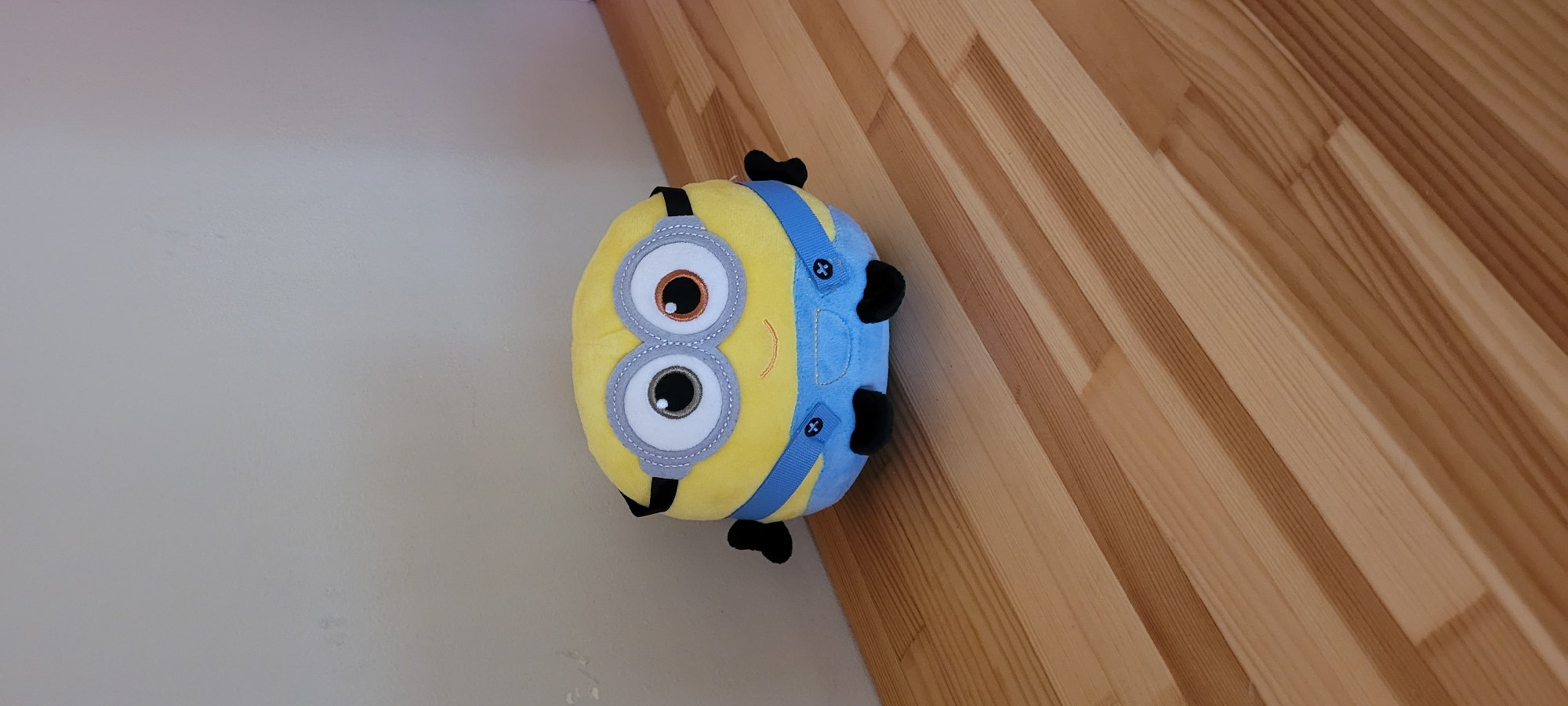 A plush toy resembling a Minion from the "Despicable Me" franchise is positioned on a light-colored wooden floor. The toy is primarily yellow with blue overalls, black shoes, and large circular goggles. The background consists of a plain, light-colored wall. The scene appears to be indoors, possibly in a residential setting. No specific location in Riga, Latvia can be identified from the image.
