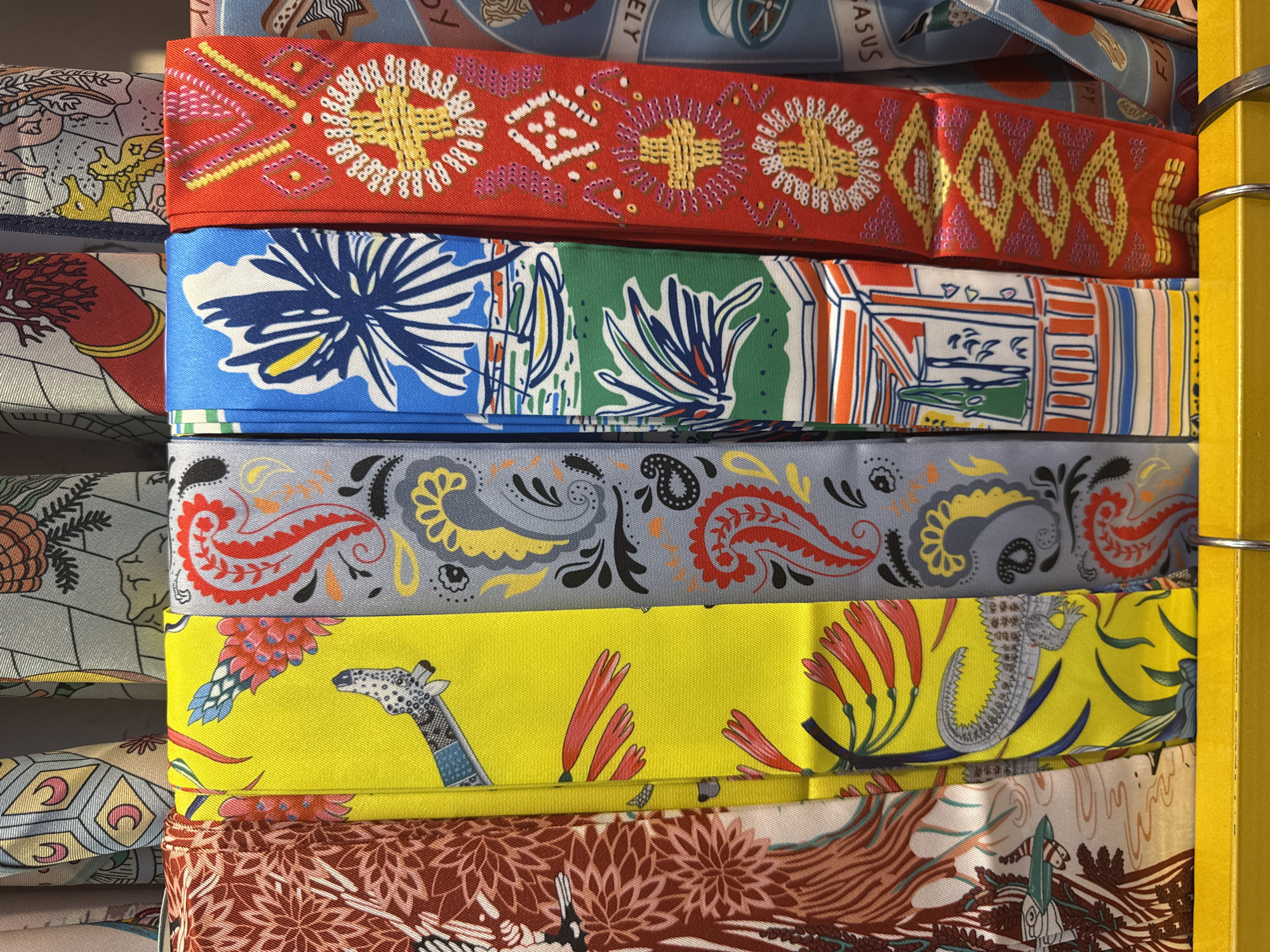 The image displays a close-up, eye-level, horizontal shot of a rack of colorful silk scarves. The scarves are folded and hung vertically, revealing a variety of intricate patterns. From left to right, the visible patterns include a dark red scarf with abstract floral motifs, a bright yellow scarf featuring a crocodile and tropical flowers, a light grey scarf with black and red paisley designs, a blue scarf depicting a beach scene with a palm tree and a sailboat, a red scarf with embroidered geometric patterns, and a light blue scarf with a detailed illustration. The scarves are displayed in what appears to be a retail setting, possibly a boutique or a market stall, with a yellow shelf visible at the bottom of the frame supporting the scarves. The lighting is even, highlighting the textures and colors of the silk. There are no people or animals present in the immediate frame, and the background is a blurred, neutral tone, suggesting an indoor environment. This scene does not depict any elements that would allow for specific localization to Nha Trang Ward, Vietnam, as the objects and environment are generic to a retail display of textiles.