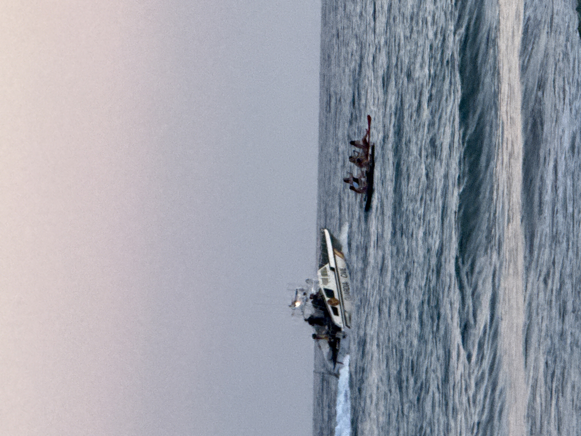 In the city of Castelldefels, Spain, on a body of water, a Guardia Civil boat with several people on board is visible on the left side of the image. The boat is white with a yellow stripe and has "GUARDIA CIVIL" printed on its side. The boat is creating a white wake. To the right of the Guardia Civil boat, there is a small raft or stand-up paddleboard with four people. The sky is a gradient of light pink and grey. The water is a dark blue with visible waves.