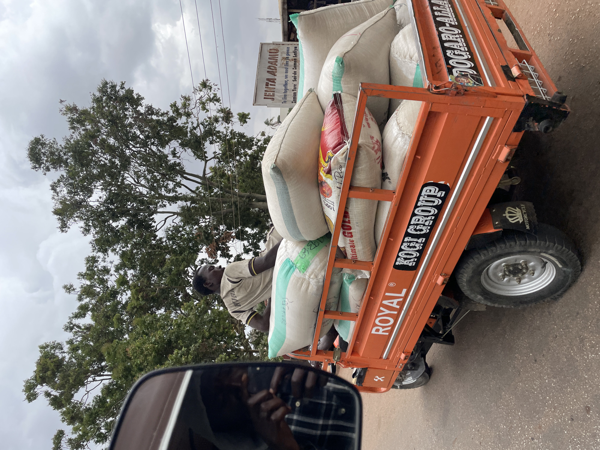 An orange three-wheeled vehicle loaded with multiple large sacks is traveling on a paved road. The sacks are mostly white, with some having colored markings. A man wearing a light-colored shirt with dark trim is driving or riding on the vehicle. The side of the vehicle has various labels including "ROYAL", "KOGI GROUP", and "DOGARO-ALLA".
In the background, a tree with green leaves stands beside the road and a sign reading "JENTA ADAMO" is visible. Utility poles with wires are positioned above the tree. The sky is overcast.
The image appears to have been taken from inside another vehicle, as a side mirror and part of the vehicle’s interior are visible in the foreground, reflecting a person wearing a plaid shirt.