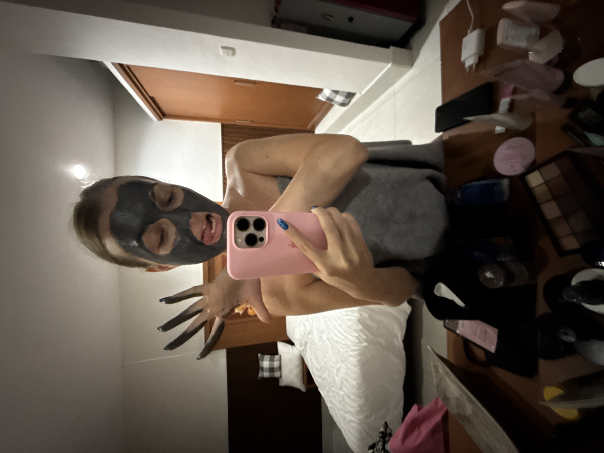 A woman with a black charcoal face mask is taking a selfie in a mirror. Her tongue is sticking out, and her right hand, with black nail polish, is raised with fingers spread. She is wearing a grey towel draped around her body. The background shows a bedroom with a white bed, a wooden headboard, and a closet. On a wooden table in the foreground, there are various cosmetics, a phone, and a charging adapter. The scene appears to be in Denpasar, Indonesia.