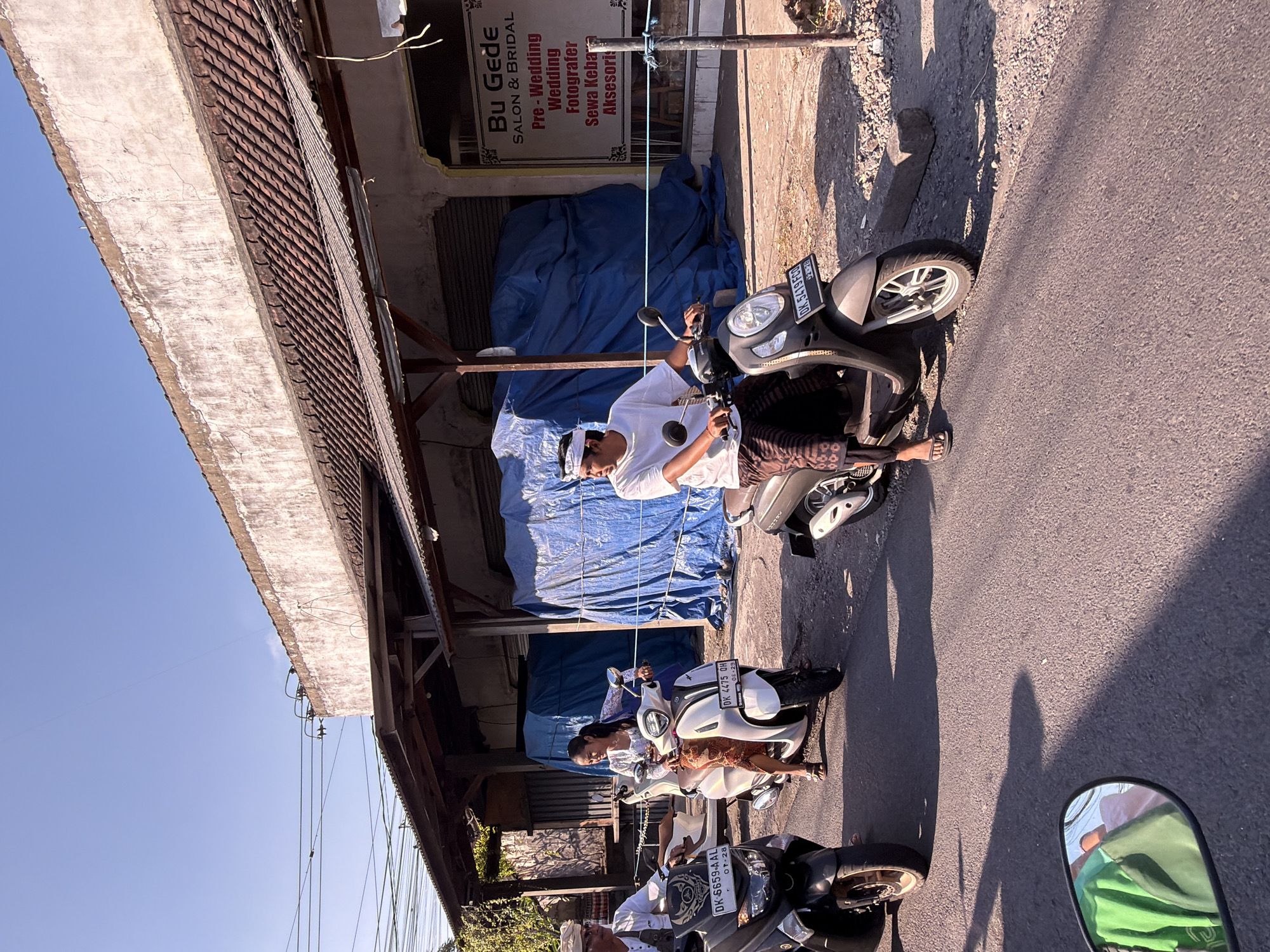 The image shows a street scene in North Kuta, Indonesia, with three people on motorbikes. In the foreground, a motorbike is partially visible with a rider wearing a green shirt, reflected in the side mirror. To the left, a man wearing a white shirt and head wrap is on a black motorbike. In the center, a woman in a white top and patterned sarong is riding a white scooter. To the right, a man in a white t-shirt and a patterned sarong is riding a dark grey motorbike. Behind them, a building with a tiled roof and weathered facade has blue tarps covering a section of its facade. A sign on the right side of the building reads "Bu Gede Salon & Bridal Pre-Wedding Wedding Fotografer Sewa Kebaya Aksesori". The road is paved, and the scene is illuminated by bright sunlight, casting long shadows. Power lines are visible overhead.