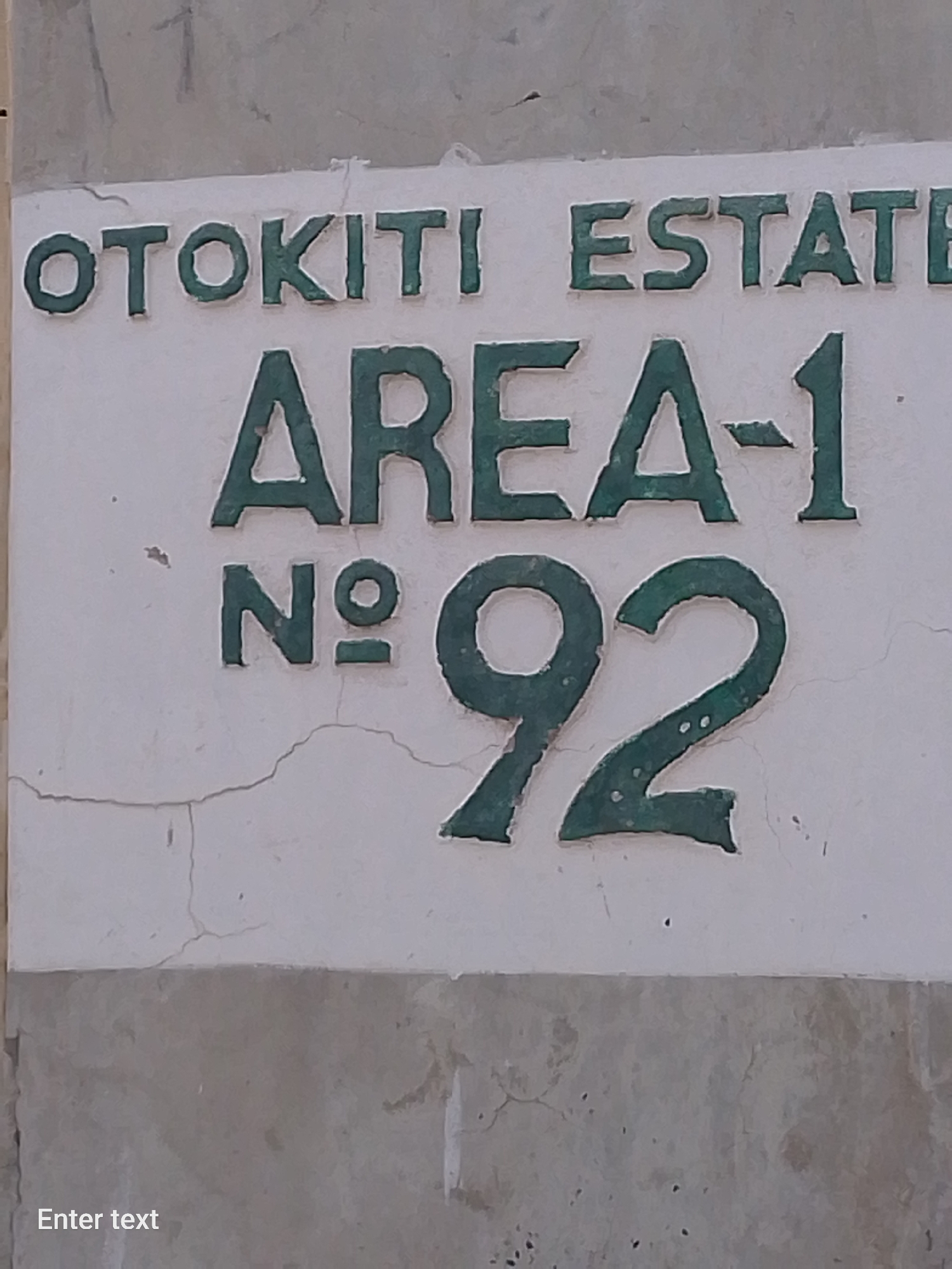 The image shows a sign that reads "OTOKITI ESTATE, AREA-1, No. 92." The sign appears to be painted in green on a white background. The background shows signs of wear and tear and is on an exterior wall. Based on this information, the location of the image may be Adankolo, Nigeria, but this can't be confirmed from the image alone. There are no people visible in the image.