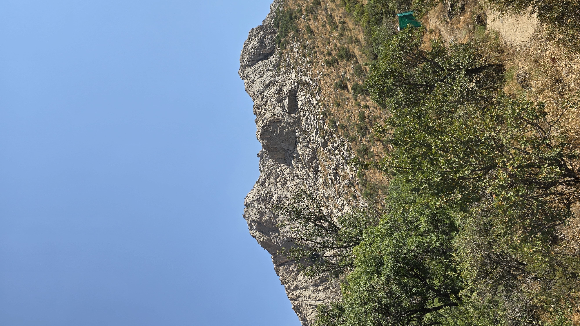 This image depicts a mountainous landscape with a clear blue sky. In the foreground, there are various types of vegetation, including trees and shrubs. A dirt path is visible, winding its way up the side of the hill. Further in the background, a large, rocky mountain rises, its slopes covered in scree and sparse vegetation. A small, bright green structure, possibly a shelter or building, is situated on the hillside. The environment appears to be dry and arid, with a sense of remoteness. The scene is devoid of any people or animals. The image suggests a natural, outdoor setting in a region with significant elevation changes.