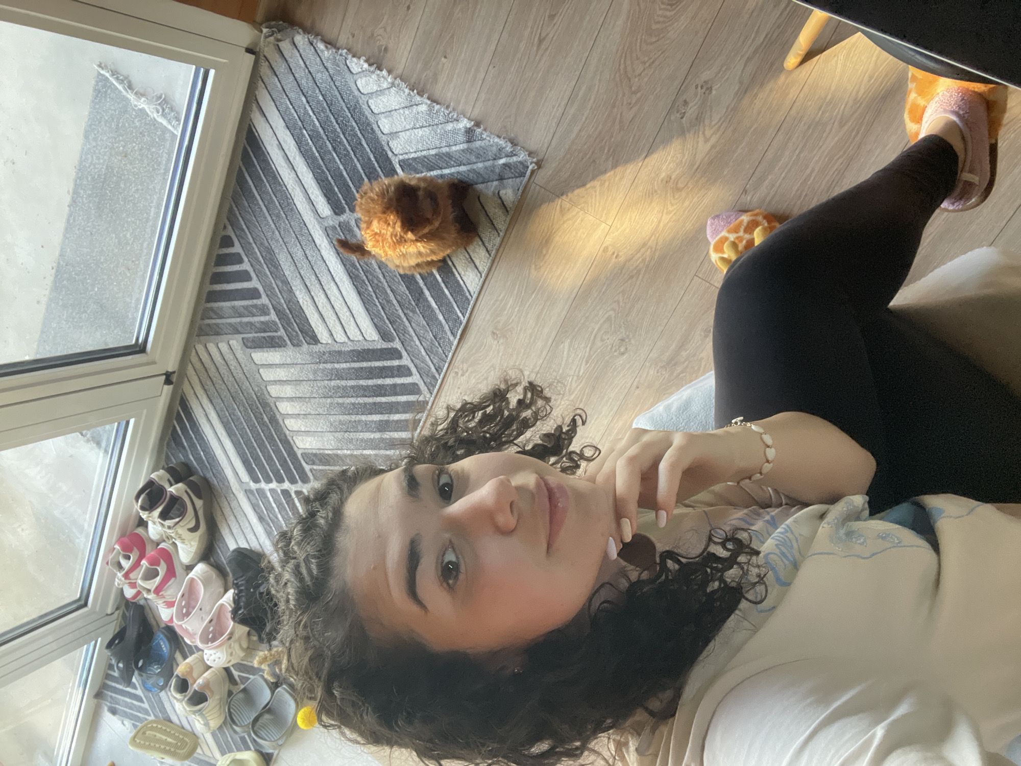 A young woman with curly dark hair is lying down, taking a selfie. She is wearing a light-colored top and black leggings. Her hand is near her chin, and she has a bracelet on her wrist. She is wearing fuzzy slippers and one that looks like a giraffe is on the floor near her feet. To her left is a sliding glass door with various pairs of shoes lined up near it. A small, fluffy brown dog is sitting on a patterned rug. The floor is light-colored wood. It's difficult to determine the exact location as the view outside does not provide many hints, but the location of the photo is said to be in Sofia, Bulgaria.