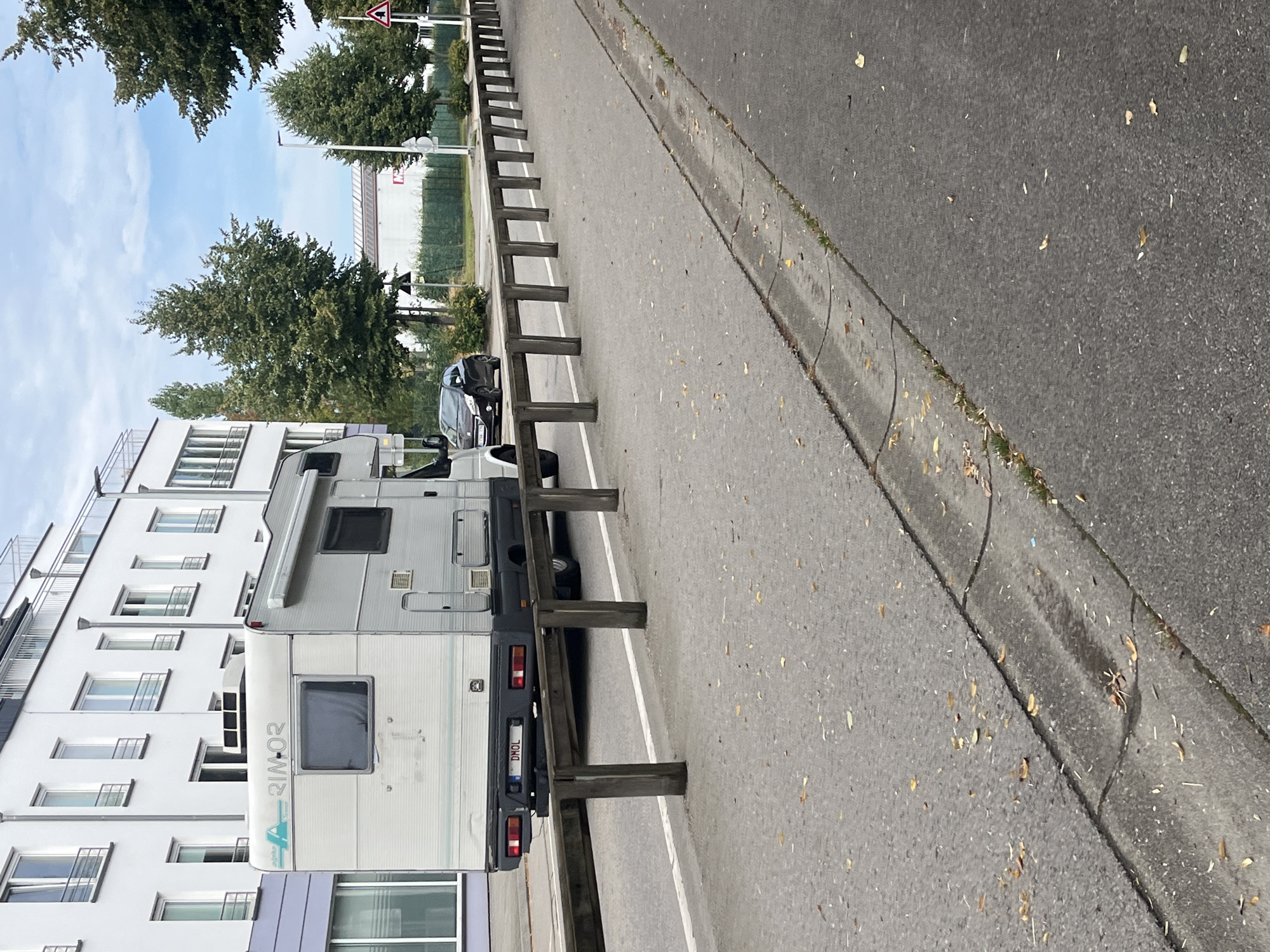 In the city of Saint-Vith, Belgium, on a cloudy day, a campervan with the license plate "DMOL" is parked alongside a street. The campervan is white and grey, with several windows and a roof-mounted air conditioning unit. It is positioned behind a wooden barrier that runs along the edge of the road. A dark-colored car is visible in the background, also parked near the campervan. The street itself is paved, with a white line marking the lane. A concrete curb and drain are visible along the side of the road, with fallen leaves scattered on the pavement and within the drain. In the background, a modern white building with multiple windows stands out, with trees and a green fence also present. A triangular warning sign is visible further down the street, indicating a sharp turn. The overall scene depicts a quiet urban street with parked vehicles and typical street infrastructure.