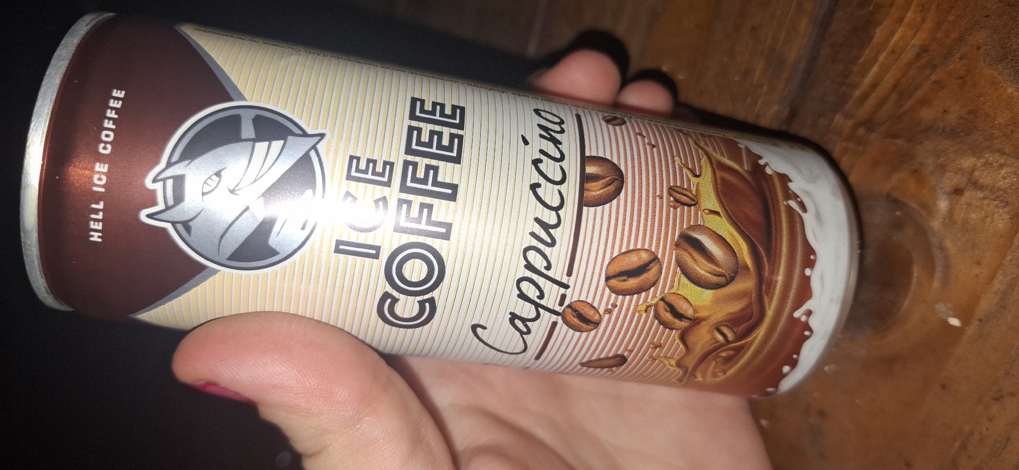 A person's hand is holding a can of "Hell Ice Coffee Cappuccino." The can is cylindrical with a brown and cream-colored design. The top of the can has the words "HELL ICE COFFEE" printed in white. Below that is a logo of a stylized devil's head. The words "ICE COFFEE" are printed in large, bold letters. Below that, "Cappuccino" is written in a script font, with coffee beans and splashes of liquid depicted. The background is dark. The can is resting on a wooden surface. The image was taken in Shumen, Bulgaria.