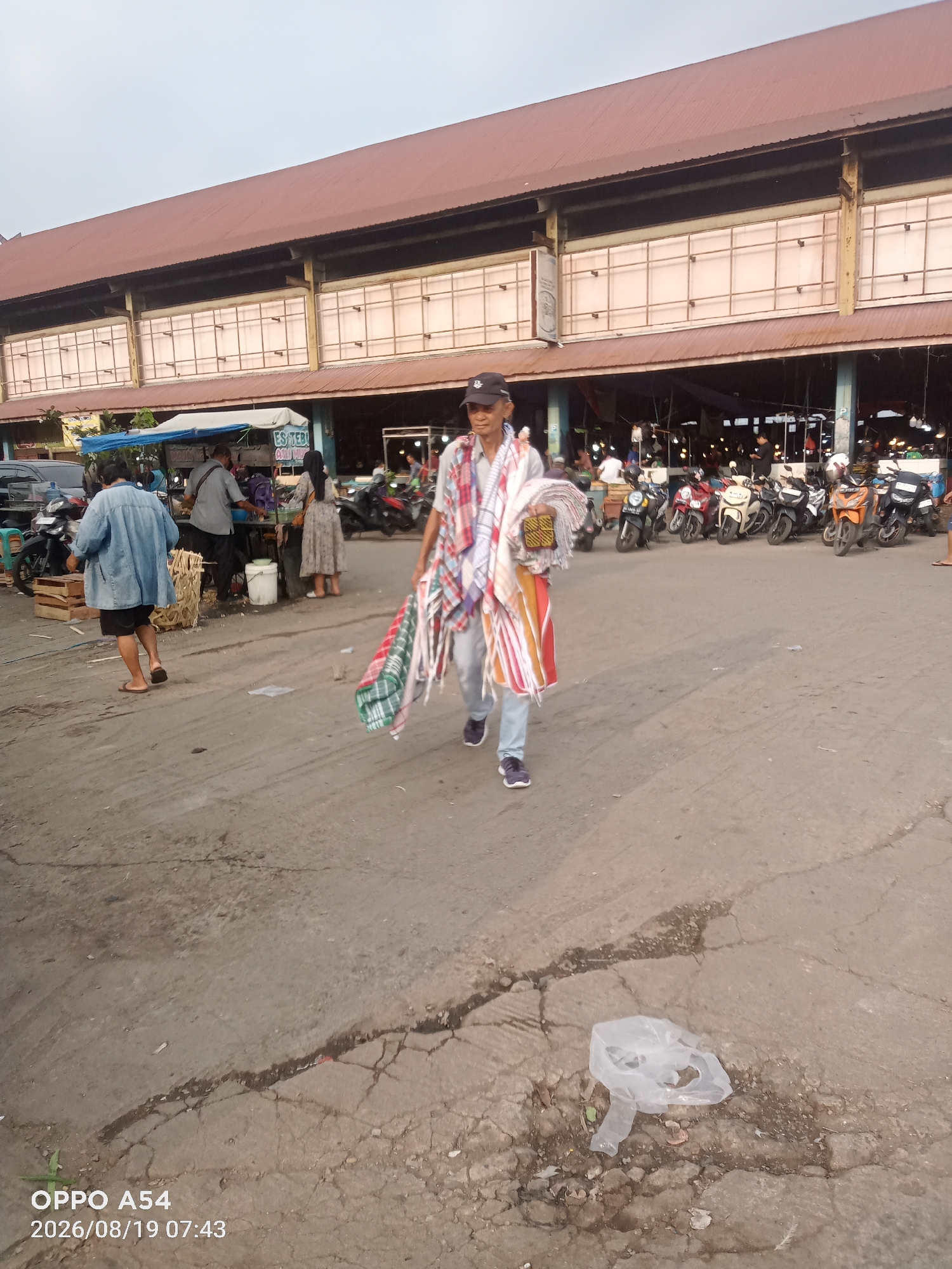 The image depicts an outdoor market scene in Jambi, Indonesia. In the foreground, a man is walking towards the viewer, carrying several folded textiles. He is wearing a light-colored shirt, grey pants, and dark sneakers, with a black cap on his head and a checkered scarf around his neck. Behind him, to the left, a woman in a denim jacket and shorts walks away from the camera. Further back, near a stall with a blue awning, several people are gathered, including a man in a grey shirt and another woman in a patterned dress. A row of motorcycles is parked in the middle ground, and in the background, a large market building with a reddish-brown roof is visible. The ground is paved with cracked concrete, and a discarded plastic bag lies on the ground in the lower right portion of the image. The time stamp on the image indicates it was taken on August 19, 2026, at 07:43.
