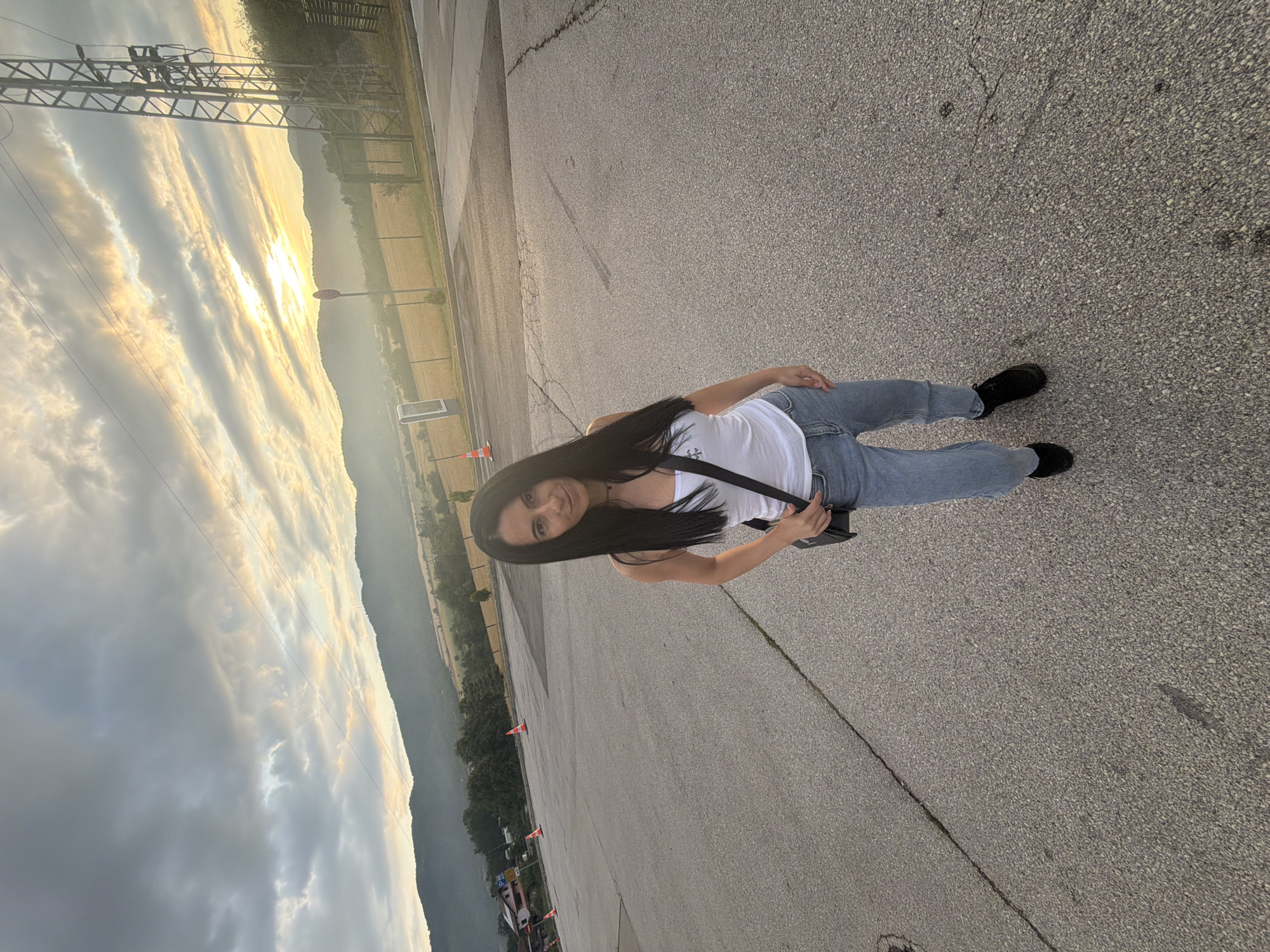 A woman with long, dark hair stands in an asphalt parking lot. She is wearing a white tank top, light blue jeans, and black shoes. She is holding a black crossbody bag. In the background, there are mountains, a field, a power line tower, traffic cones, and a sign. The sun is setting, casting a warm glow over the scene. The image appears to have been taken in Razliv, Bulgaria, based on the description provided.