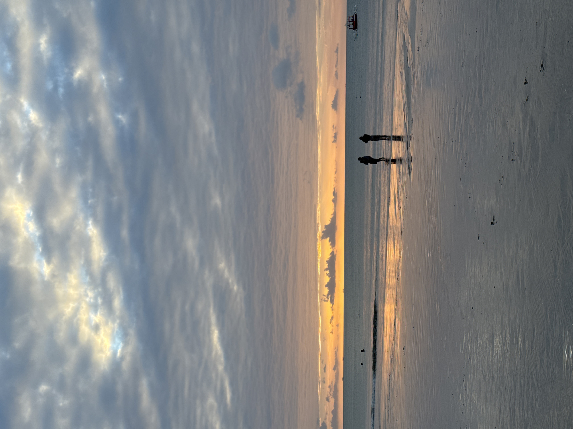 The image depicts a serene beach scene at sunset in Kuta Selatan, Indonesia. The sky is a canvas of soft, muted colors, with streaks of yellow and orange from the setting sun peeking through a blanket of gray clouds. The ocean stretches out, calm and reflective, mirroring the sky's gentle hues. A sandy shore, wet from the receding tide, lies in the foreground, marked by subtle ripples and scattered debris.
Two individuals stand near the water's edge, their silhouettes dark against the luminous horizon. One person appears to be holding something, possibly a child or a bag, while the other stands beside them, both seemingly engaged in a quiet observation of the natural spectacle. Further out in the water, several other figures are barely visible, suggesting others are also enjoying the beach. A small, red boat with a distinctive roof is visible in the distance, adding a touch of man-made presence to the otherwise untouched landscape. The overall atmosphere is one of peaceful solitude and appreciation for the beauty of nature.