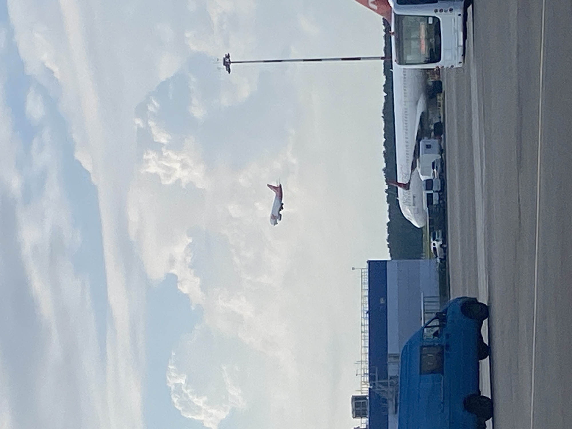 A passenger airplane, predominantly white with red tail fins, is ascending into a partly cloudy sky over an airport tarmac. In the foreground, a blue panel van is parked on the tarmac. Behind the van, a long white airplane is stationary on the ground. To the right of the stationary airplane, a white bus is visible, and further right, a red and white striped airport light pole stands tall. In the background, a long, blue and grey building is situated behind the blue van, with a dense green treeline on the far horizon. The scene appears to be at Pulkovo Airport in Saint Petersburg, Russia.