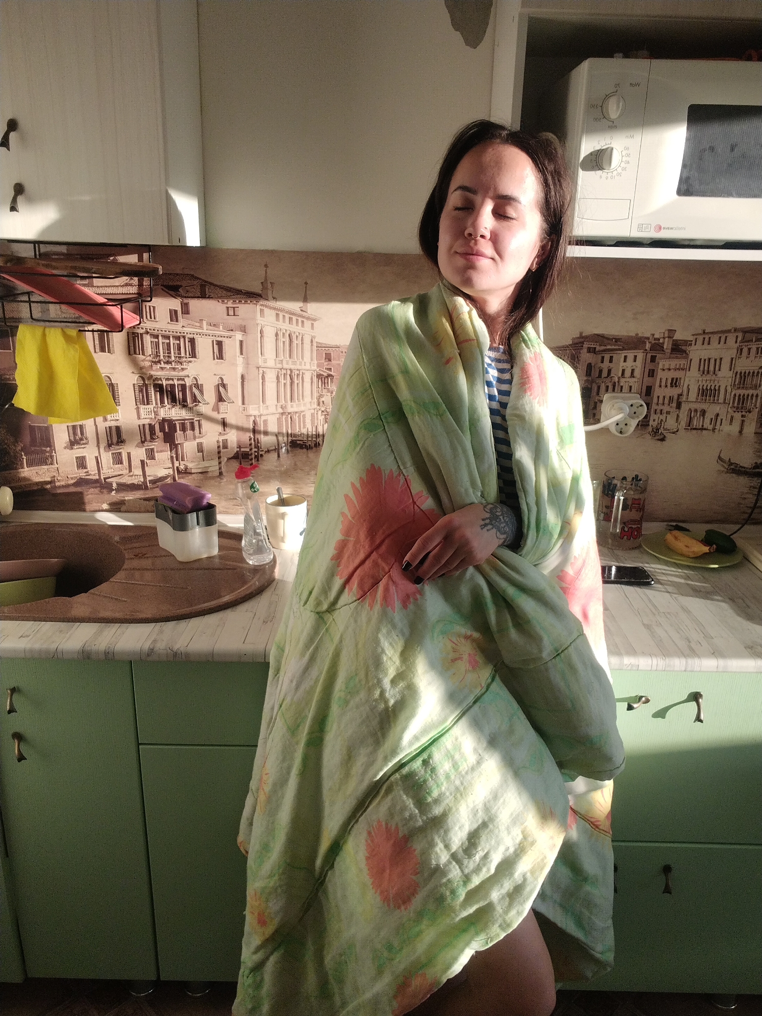 A young woman stands in a kitchen, her eyes closed in serene enjoyment as warm, golden sunlight illuminates her face. She has dark hair and is wrapped in a light-colored, possibly linen, blanket or throw adorned with large orange/pink and yellow floral patterns. A striped top is visible underneath the blanket, and a tattoo is seen on her left forearm.
The kitchen features light green base cabinets with brass-colored handles and a light-toned countertop with a wood-grain pattern. On the left, a dark brown, circular sink is integrated into the counter. Nearby, a black metal dish rack holds a yellow cloth and a pink drying mat. A clear dish soap dispenser and a grey and white sponge holder with a purple sponge are also on the counter. Further along, a yellow mug sits, and a white microwave oven with the text "evrocuisine" is positioned on a shelf above. On the right side of the counter, a clear glass with "HO" visible text, a green plate holding a banana peel and a green vegetable, and a dark smartphone are present. A white wall-mounted power strip is also visible on the wall.
The background wall is covered with wallpaper or a mural depicting a Venetian-style cityscape, featuring old buildings and canals. The strong, low-angle sunlight suggests it is either early morning or late afternoon, creating a peaceful and comfortable atmosphere. The woman's relaxed posture and closed eyes convey a moment of contentment and warmth in what appears to be a home kitchen in Noviy, Russia.