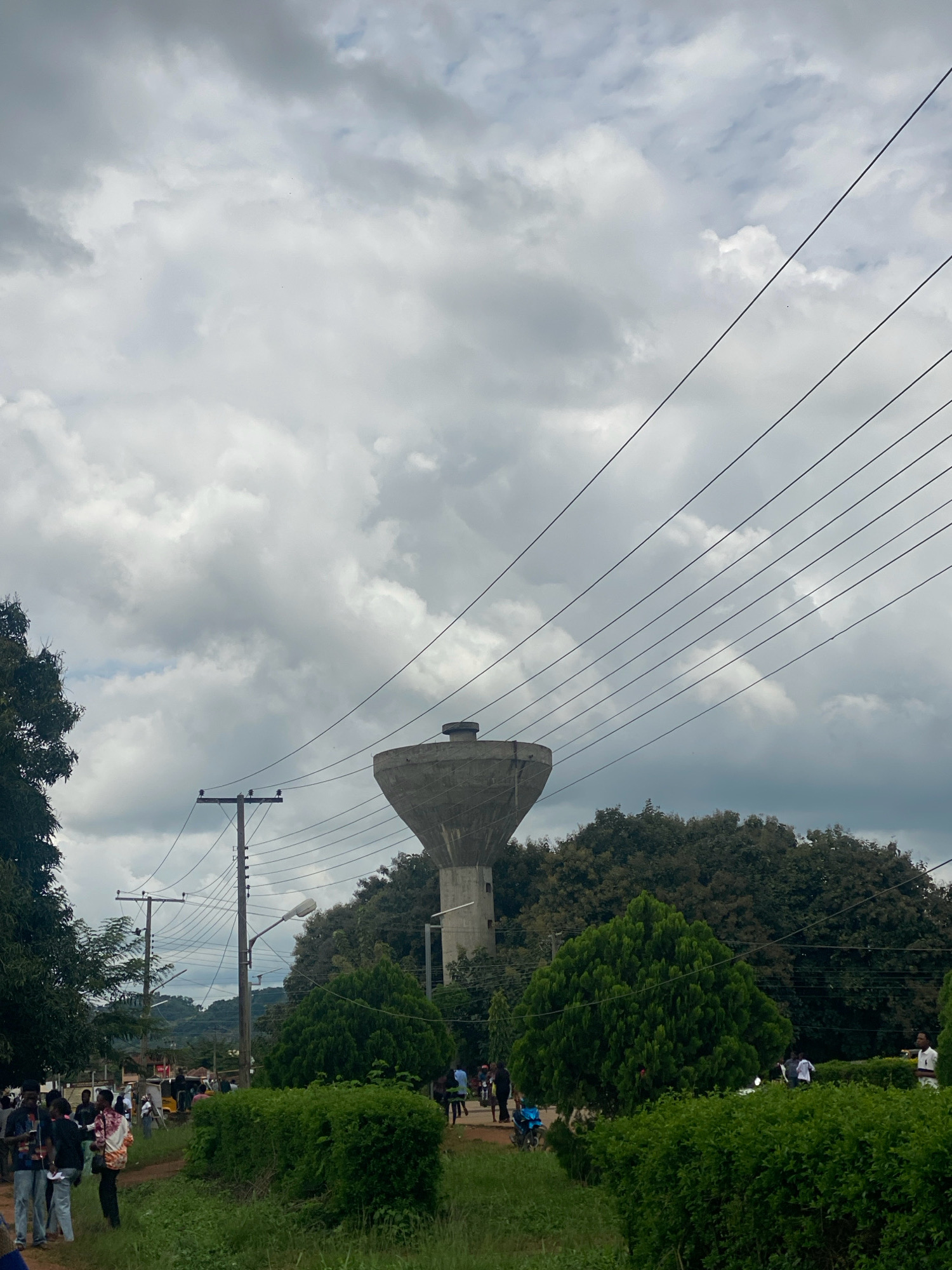 This is a medium shot, exterior, daytime photograph taken in Iworoko, Nigeria. The scene is dominated by a large, concrete water tower, visible in the upper-middle section of the frame. The tower has a wide, bowl-like top and a cylindrical base. It is surrounded by lush, dense green trees. In the foreground, there are several people walking and standing, some near a dirt path. They are casually dressed. There are also several utility poles with wires crisscrossing the sky. The sky is overcast with heavy, gray clouds. The overall environment appears to be a public space or park area with some infrastructure.