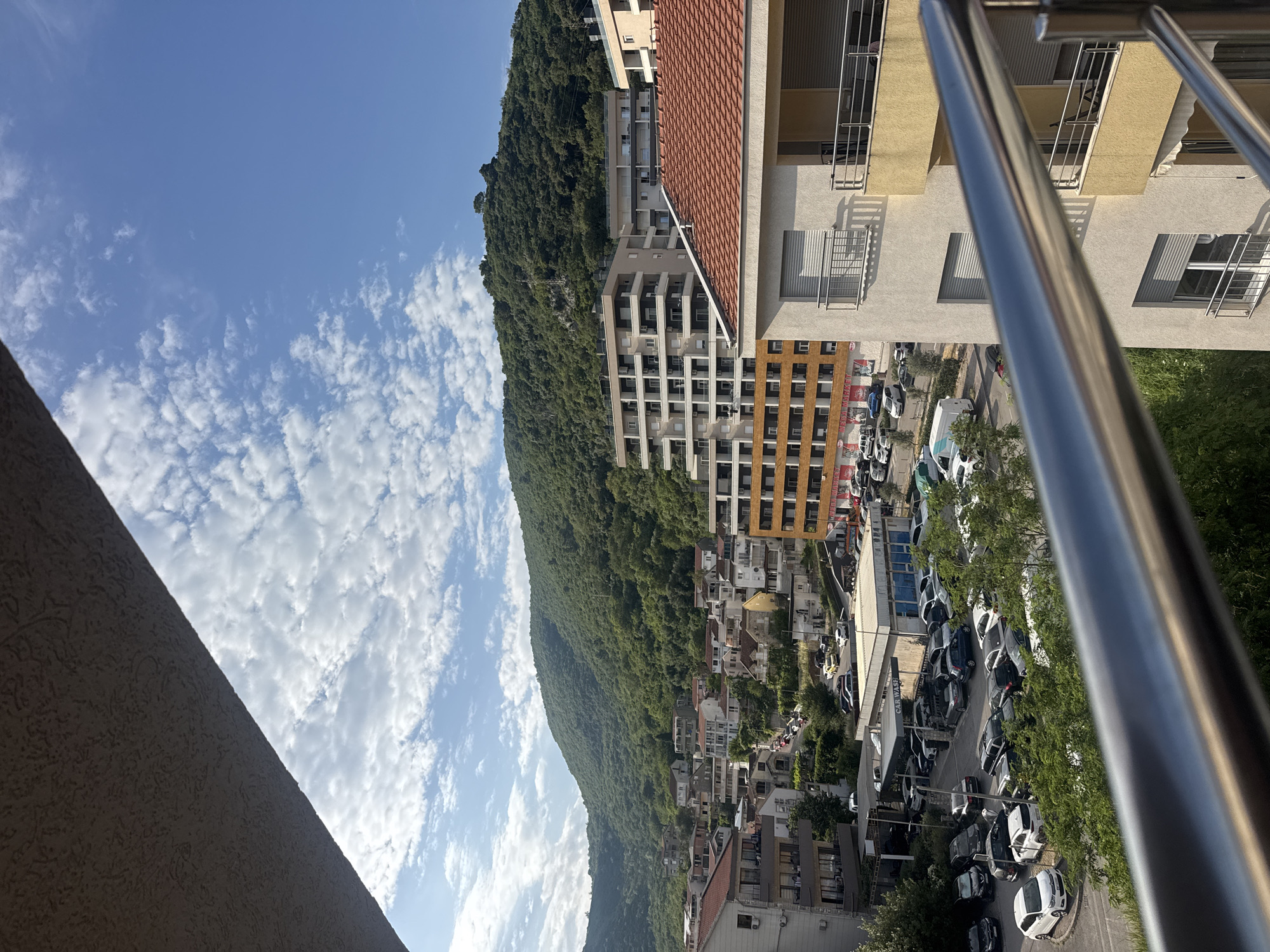 The image captures a view of a residential area in Budva, Montenegro, dominated by a verdant hillside covered in dense trees. In the foreground, a metal railing of a balcony is visible, with a textured ceiling above it. Below the balcony, a cluster of buildings, some modern with multiple stories and balconies, and others older with red-tiled roofs, line the streets. A significant portion of the scene is occupied by a parking lot filled with various cars, and a car wash facility with blue siding. The sky is bright blue with scattered white clouds, indicating a clear day. The overall impression is a bustling urban environment nestled against a natural, mountainous backdrop.