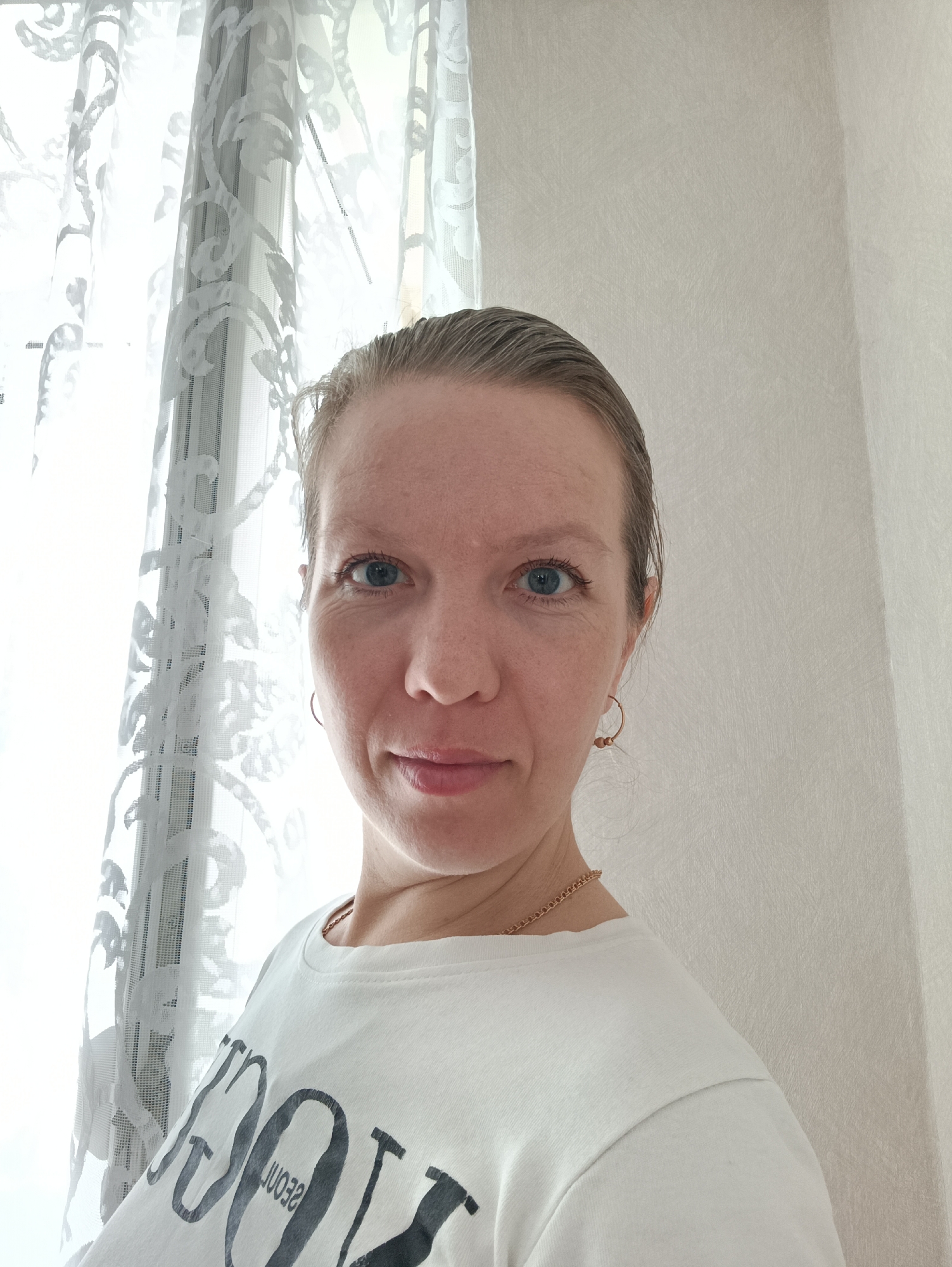A woman with fair skin, blue eyes, and her hair pulled back is in a room with white walls. She is wearing a white t-shirt and gold hoop earrings. A gold necklace is visible. She is looking at the camera with a slight smile. To her left is a window with a white, patterned curtain. Based on the context of being in the city of Chaikovsky, Russia, the environment is likely a residential interior.