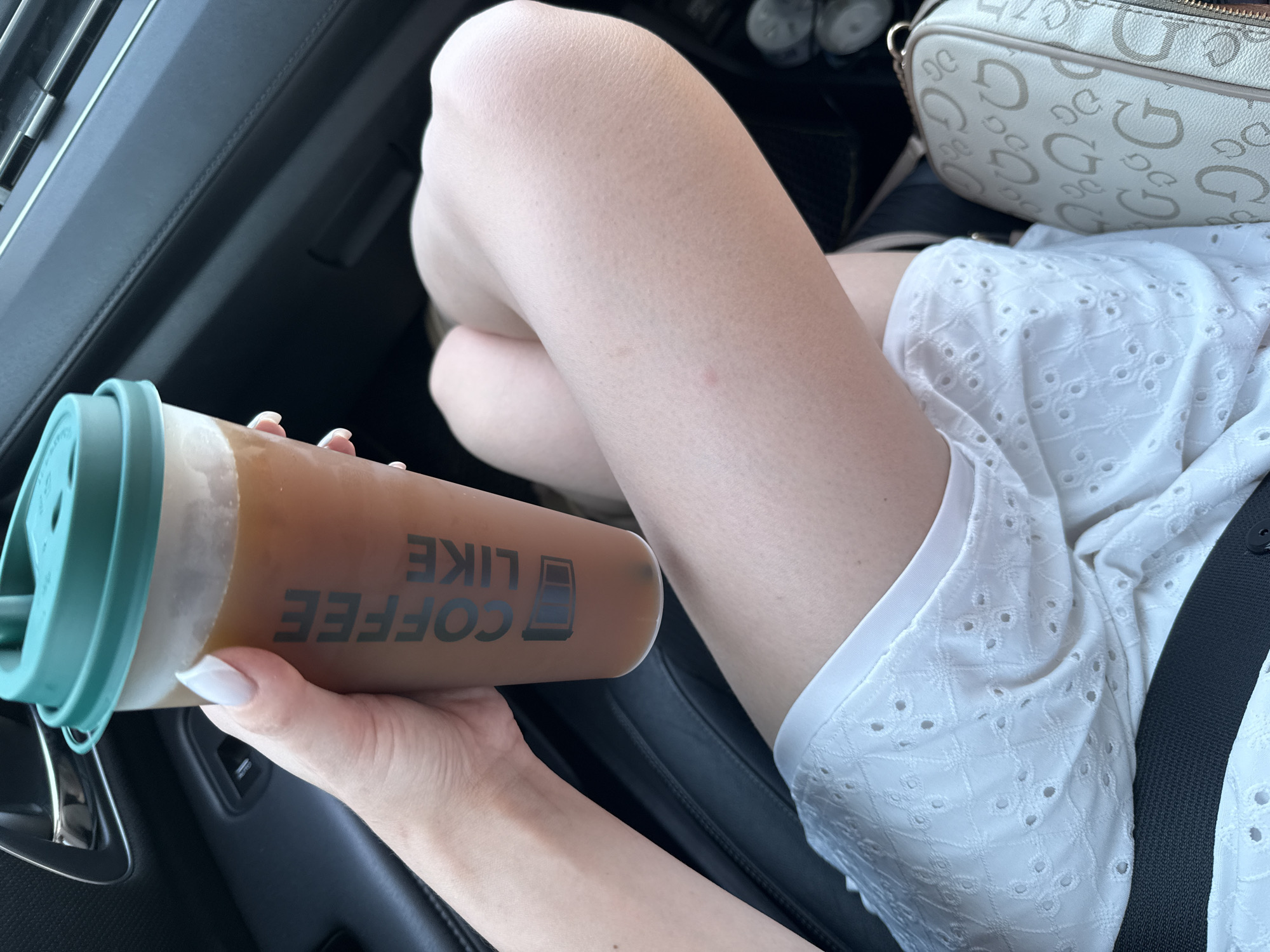 A woman sits in the passenger seat of a car holding a to-go coffee cup. Her right hand, adorned with white-tipped fingernails, grips the cup which has "COFFEE LIKE" printed vertically on its side in a dark font. She is wearing a white eyelet skirt. Next to her is a cream-colored crossbody bag with a repeating "G" logo. The interior of the car is dark, with black leather seats visible. The setting is indoors, in a vehicle.