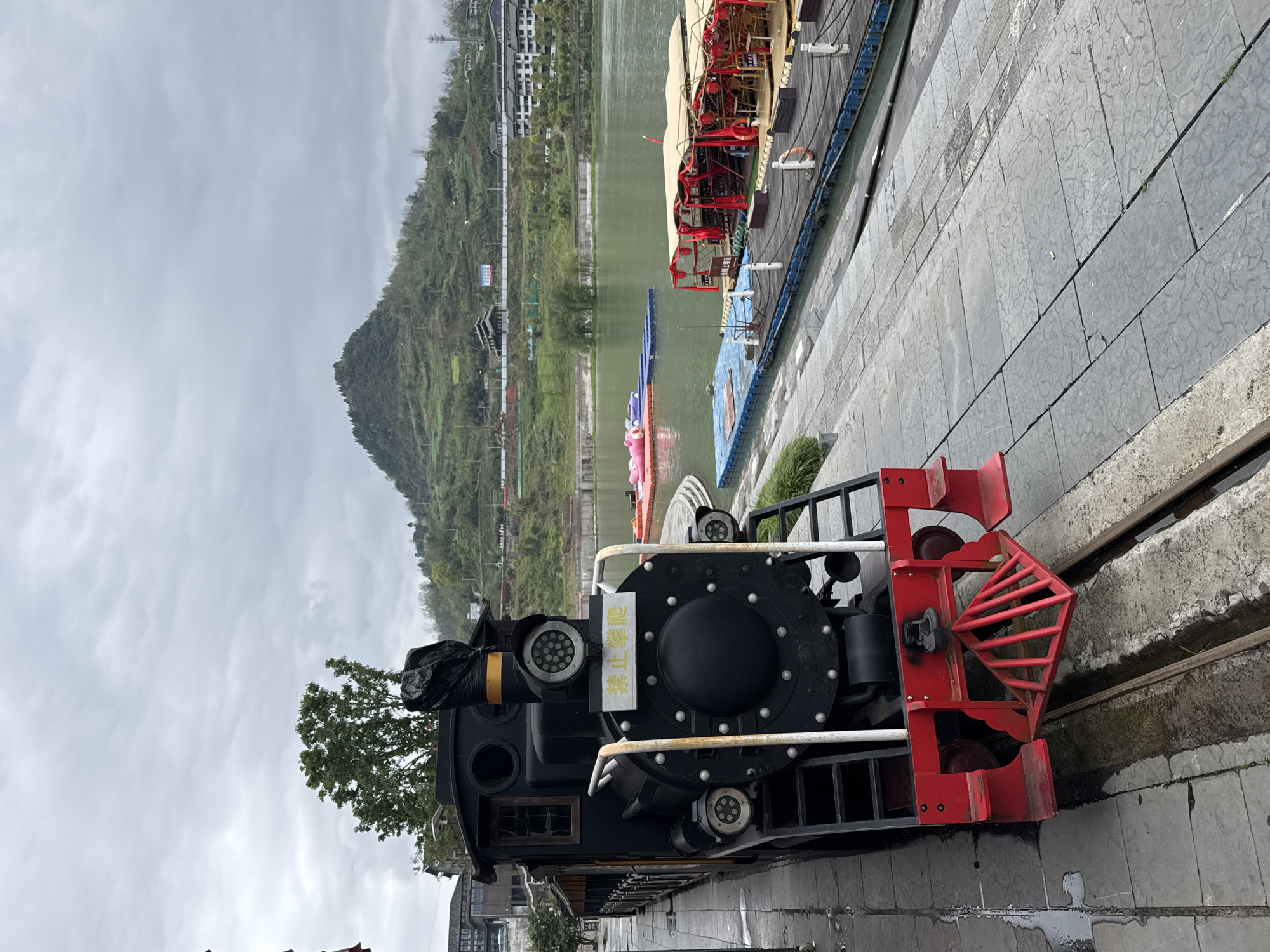The image shows a black steam train replica with a red front grill, situated on a section of track on a paved surface. The train is facing right, and a tree grows behind the locomotive. A body of water is in the background, with boats docked along a pier. The surrounding area includes buildings on a hillside and a large, green mountain under a cloudy sky. The location is Zhuoshui, China.