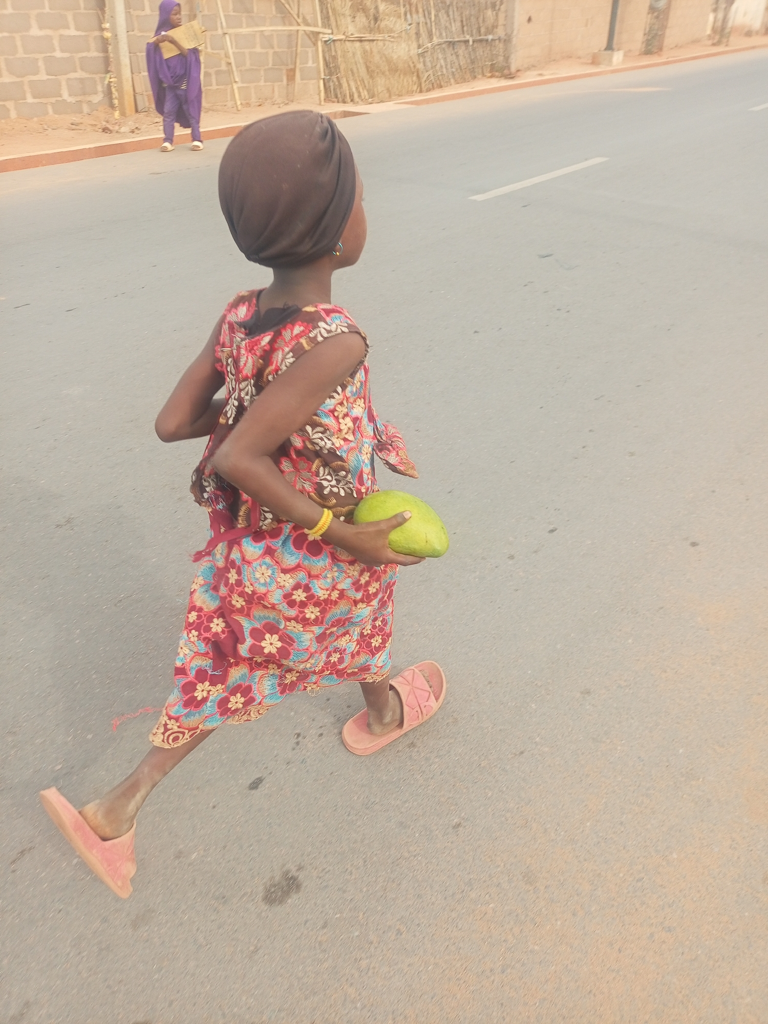 A young person, likely a child, is captured from behind, walking on a light gray asphalt road in Tempure, Nigeria. The child wears a vibrant, multicolored sleeveless dress with patterns of red, blue, and gold, and a plain brown head covering resembling a turban. They have a yellow bracelet on their left wrist and a small hoop earring on their right ear. In their left hand, they hold a large, unripe green fruit, possibly a mango. The child is wearing pink slip-on sandals, with the left sandal appearing slightly askew.
Further down the road, in the background, another individual dressed in a purple garment that covers their head and body is visible, walking or standing near a sand-colored block wall. Behind this wall, there are some rough wooden structures. The road itself is mostly clear, with a faint white line visible in the distance. The overall lighting suggests it is daytime, possibly early morning or late afternoon, given the soft, warm tones in the scene. The weather appears clear. No text is visible in the image.