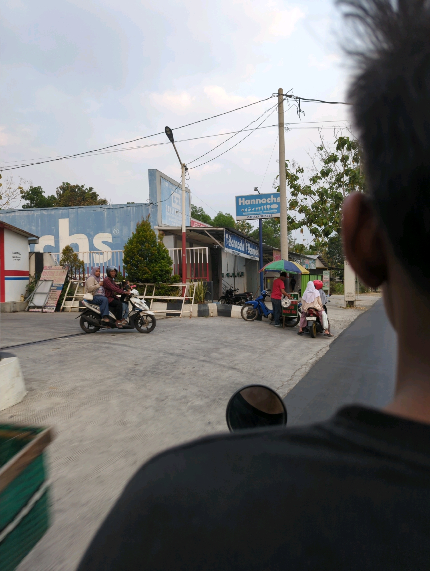 The image captures a street scene in Senggrong, Indonesia. In the foreground, the back of a person's head and shoulder are visible, suggesting the photo was taken from their perspective, possibly from a moving vehicle. The background features a light blue building with large white lettering that appears to be "cbs". To the right of this building, a pole with electrical wires and a street lamp is visible, along with a blue sign that reads "Hannochs" and "CAHAYA SUKSES". Several people are present. On the left, two individuals are riding a white motorcycle. In the middle of the frame, a man in a red shirt is interacting with two people on a scooter. One of them is wearing a red hijab. A blue motorcycle is parked nearby. The ground is paved, and there are some trees and foliage visible, along with a green bin in the lower left corner. The sky is cloudy.
