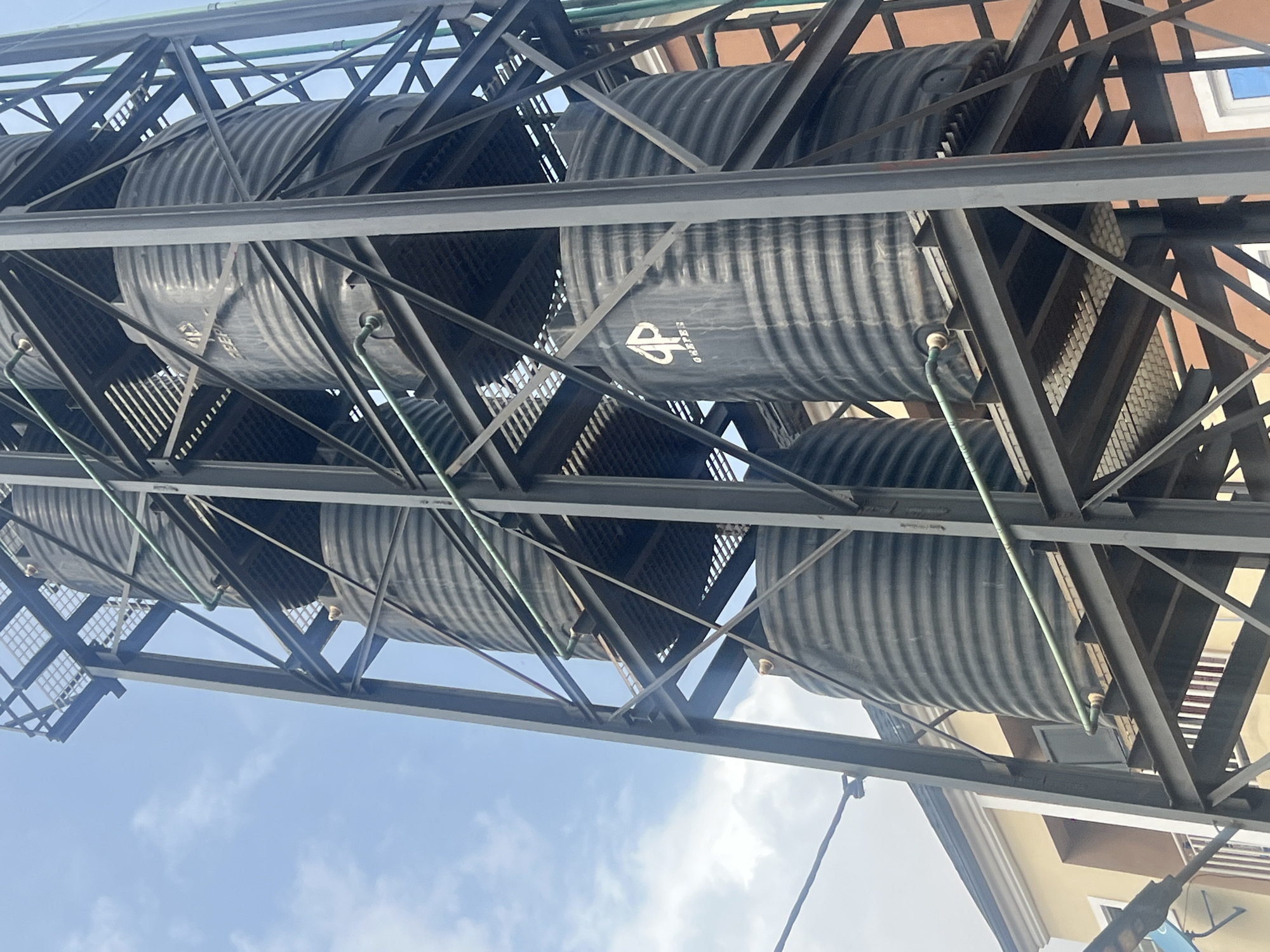 The image shows multiple black water tanks stacked on a steel framework in Egan, Nigeria. The tanks are cylindrical with corrugated exteriors and markings, including a visible "GP" logo on one. The structure supporting the tanks is constructed of steel beams and gridded metal platforms. A light blue sky is partially visible. A building with beige walls is also visible in the background. A green pipe is running along the side of the tank.