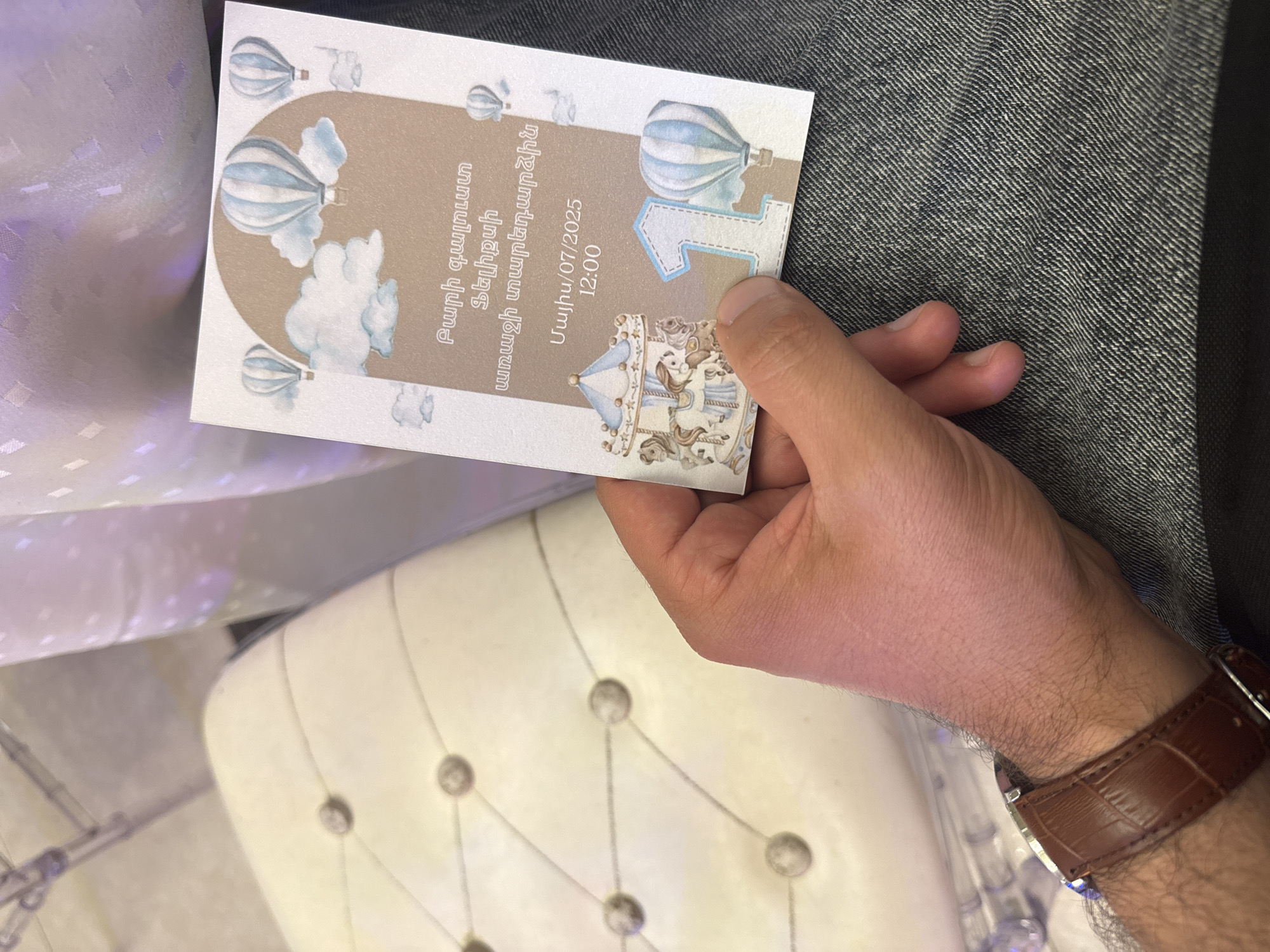 A person is holding a party invitation card. The card features images of hot air balloons and a carousel, with text in an unfamiliar script. The card indicates the date of the event as July 7, 2025, at 12:00. The person is wearing a brown leather wristwatch. The background includes a gray fabric and a white buttoned chair. The city and specific location are not identifiable in the image.