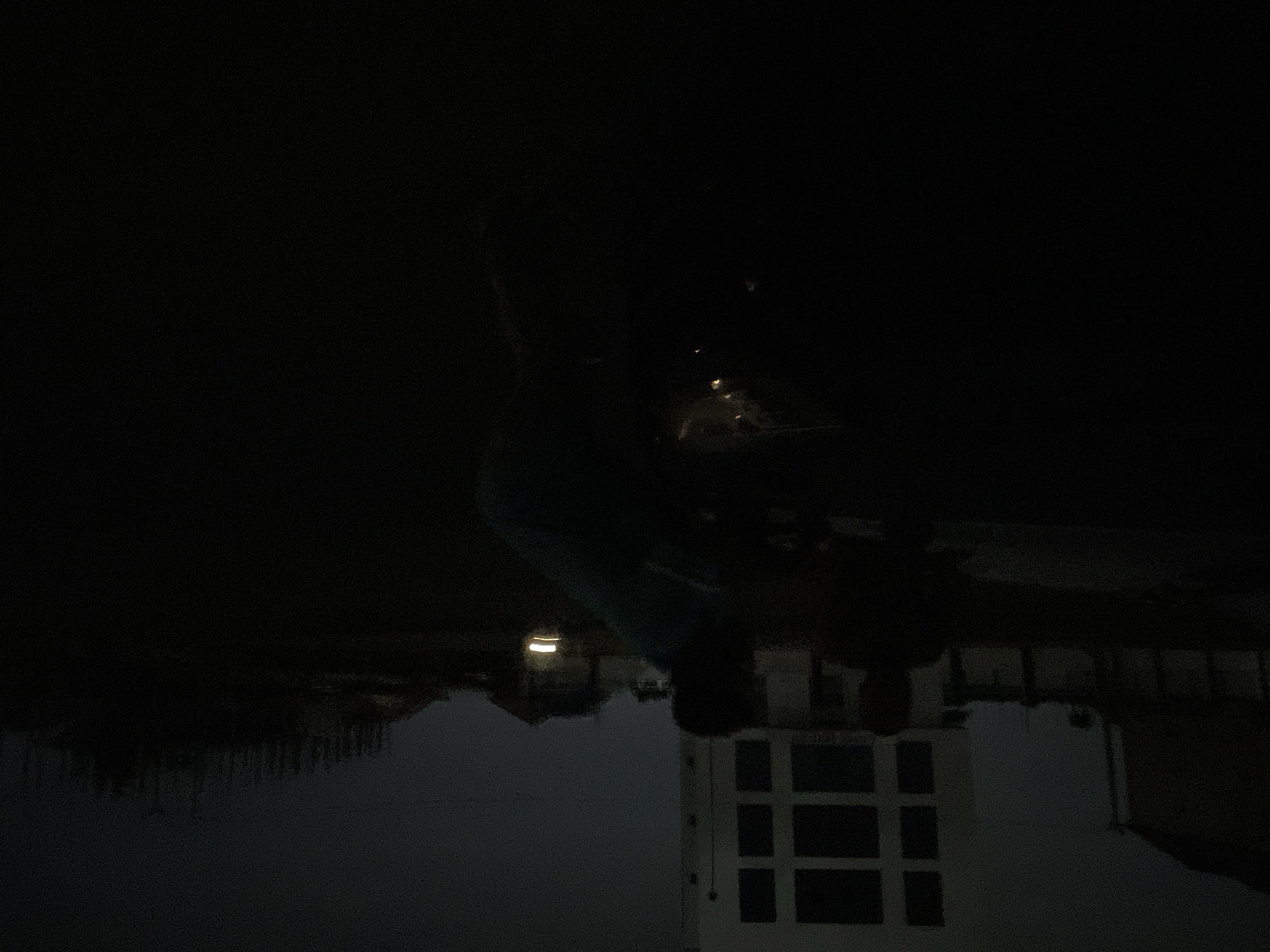 The image shows a night scene in Dutsen Alhaji, Nigeria. A white building with a grid of windows is visible in the foreground. Other structures, possibly houses, are present in the midground. Above, in the darkness, a person is visible, seemingly looking down at the scene.