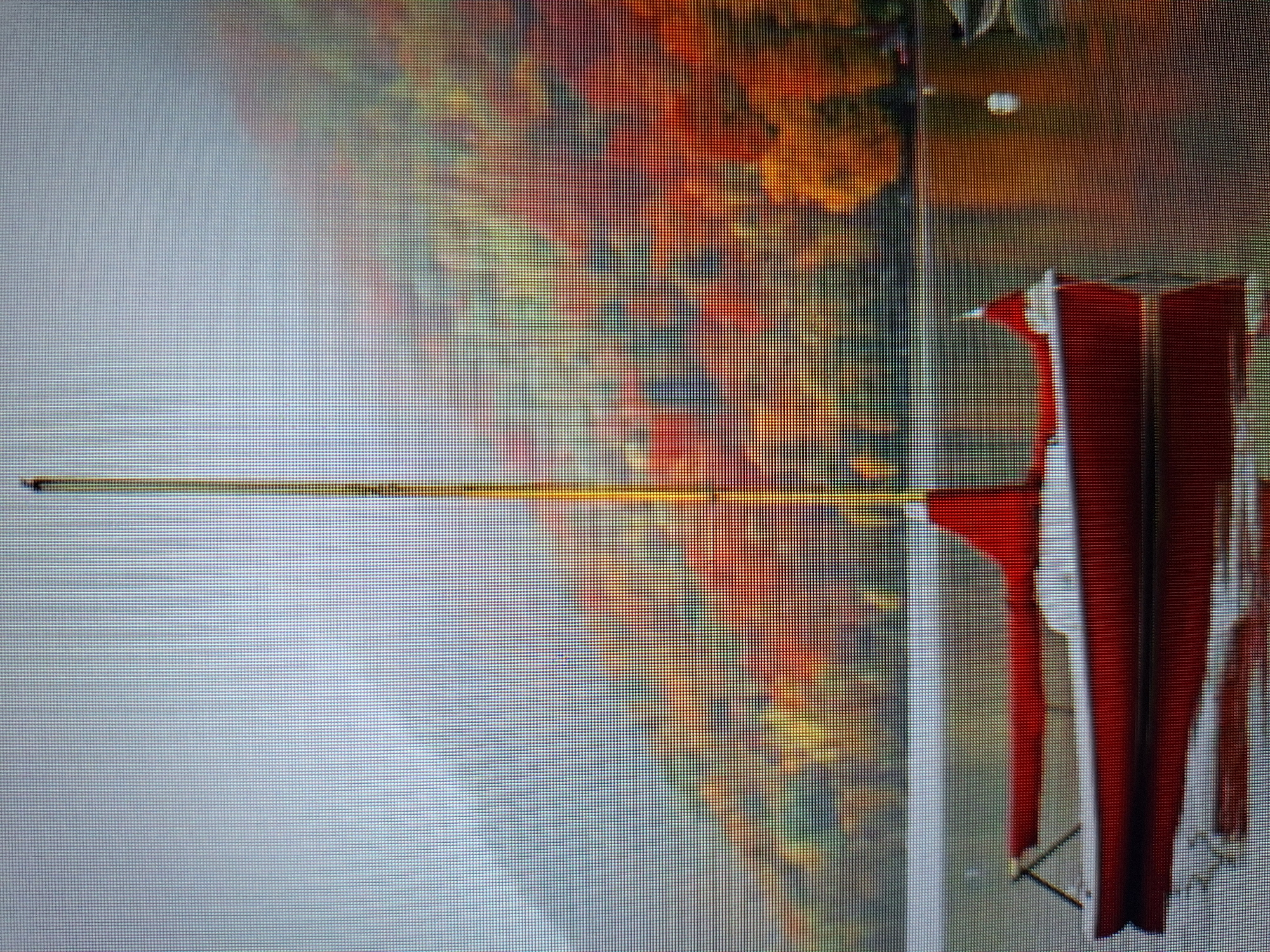 The image depicts a red and white sailboat anchored in a calm body of water. The sailboat has a tall, thin mast and a red sail that is partially furled. The hull of the boat is red, and the deck appears to be white. The boat is reflected in the water below. In the background, a lush hillside is covered in trees displaying vibrant autumn foliage in shades of red, orange, and yellow. The sky is overcast and appears misty or foggy, obscuring the distant mountain range. The overall scene suggests a serene, autumnal day in a natural setting. No specific location in the city of Varna, Bulgaria, can be identified from the provided image.