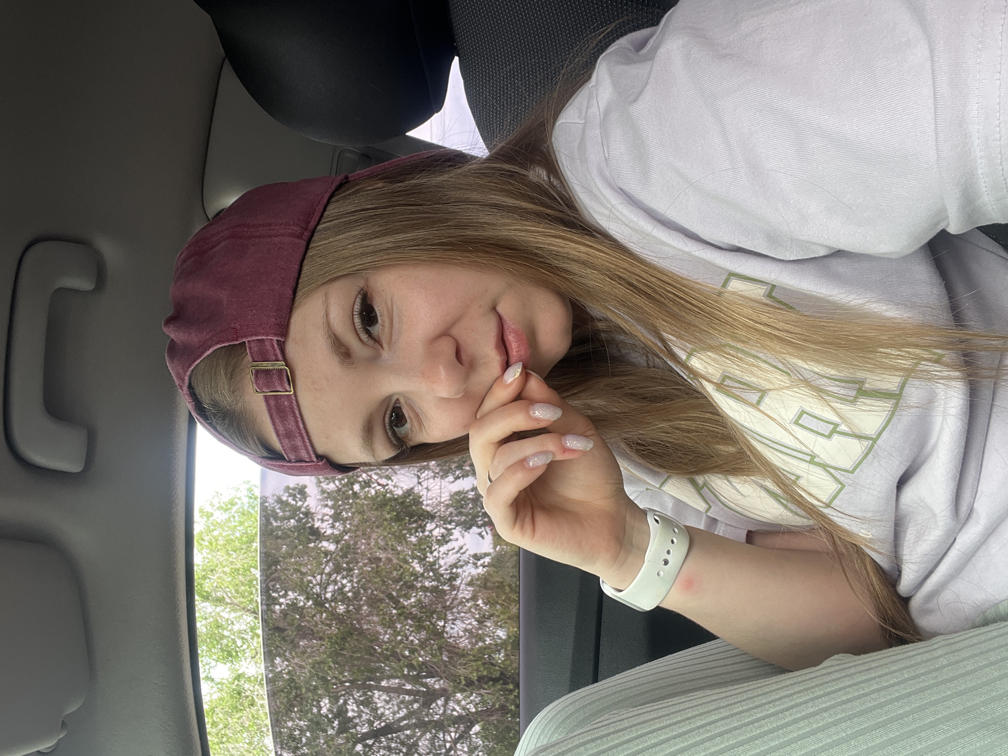 A young woman with long blonde hair, wearing a maroon baseball cap and a light purple t-shirt, sits in the passenger seat of a car. She is looking towards the camera with her hand near her mouth, and her nails are painted with glitter. A white smartwatch is visible on her left wrist. Outside the car window, trees and foliage are visible, suggesting an outdoor setting. The interior of the car shows a black headrest and a gray ceiling handle. No specific landmarks in Shadrinsk, Russia, can be identified from the image.