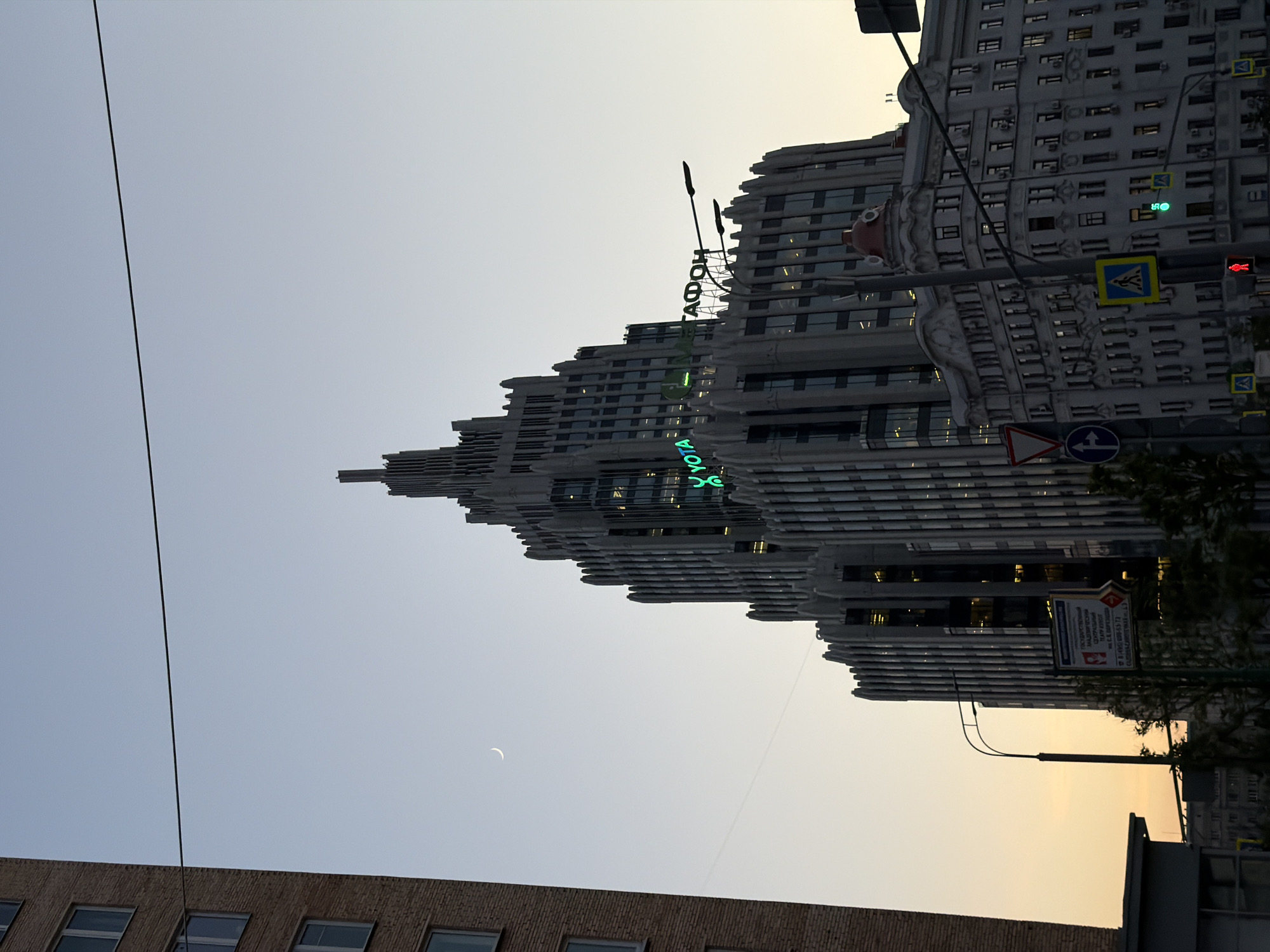 The image captures a vertically oriented street scene in Moscow, Russia, at dusk. The dominant feature is a large, multi-tiered building with prominent architectural elements and a neon sign reading "SOKOL". To the right of this modern structure, there is an older, more traditionally styled building, complete with intricate detailing and architectural features. Street signs are visible. In the foreground, a lower building is visible. A crescent moon hangs in the sky, contributing to the overall composition. A wire runs across the sky. The colors in the sky transition from a light blue to a golden hue near the horizon, suggesting the sun has recently set. The scene is a mix of architectural styles.
