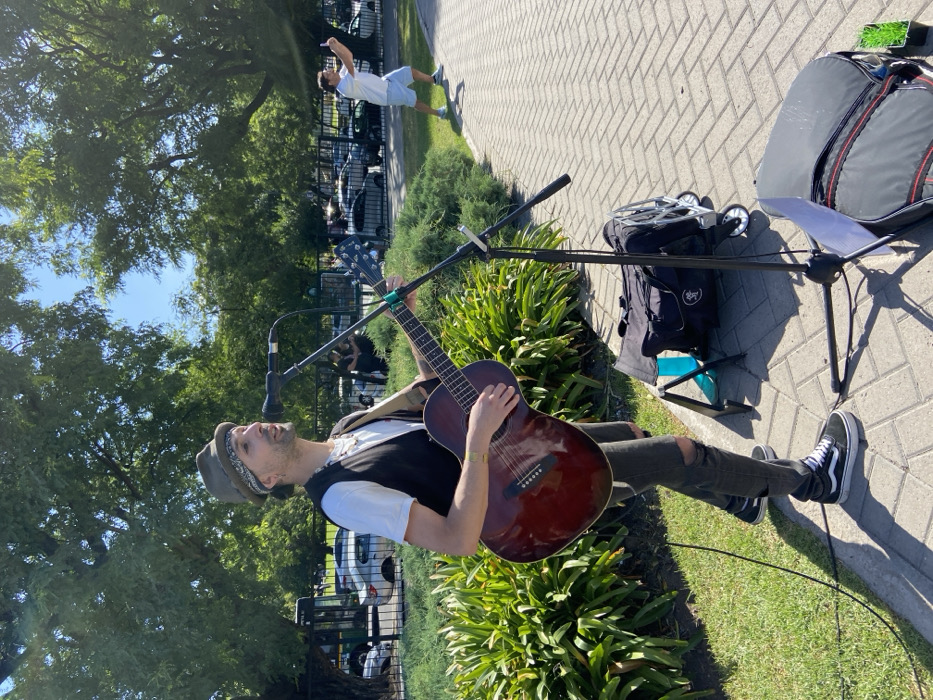 A musician in a hat, white shirt, black vest, and ripped jeans plays guitar and sings into a microphone outdoors. He sits on a small stool between leafy bushes, with a black rolling backpack and sheet music on a stand nearby. A person in a white shirt and light shorts stands and gestures in the background on a brick path, lined with trees and cars.
