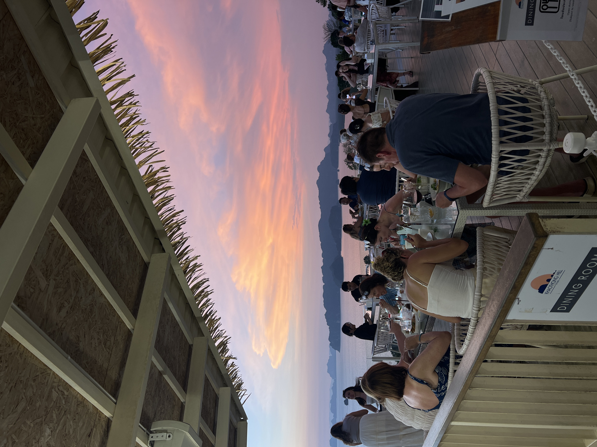 Here is a description of the image:
The photograph captures a group of people dining at what appears to be an outdoor restaurant in Khao Thong, Thailand, as indicated by the sign that reads "KHAOTHONG HILL DINING ROOM". It is evening, and the sky is a vibrant mix of pink, orange, and purple hues, suggesting a sunset. The restaurant has a thatched roof visible on the left side of the frame. Diners are seated at tables, engaged in conversation and eating. The environment includes a body of water in the background, likely part of the coastal area. The furniture is mostly white wicker. Some diners are dressed casually. A printed sign is visible that advertises dining availability.