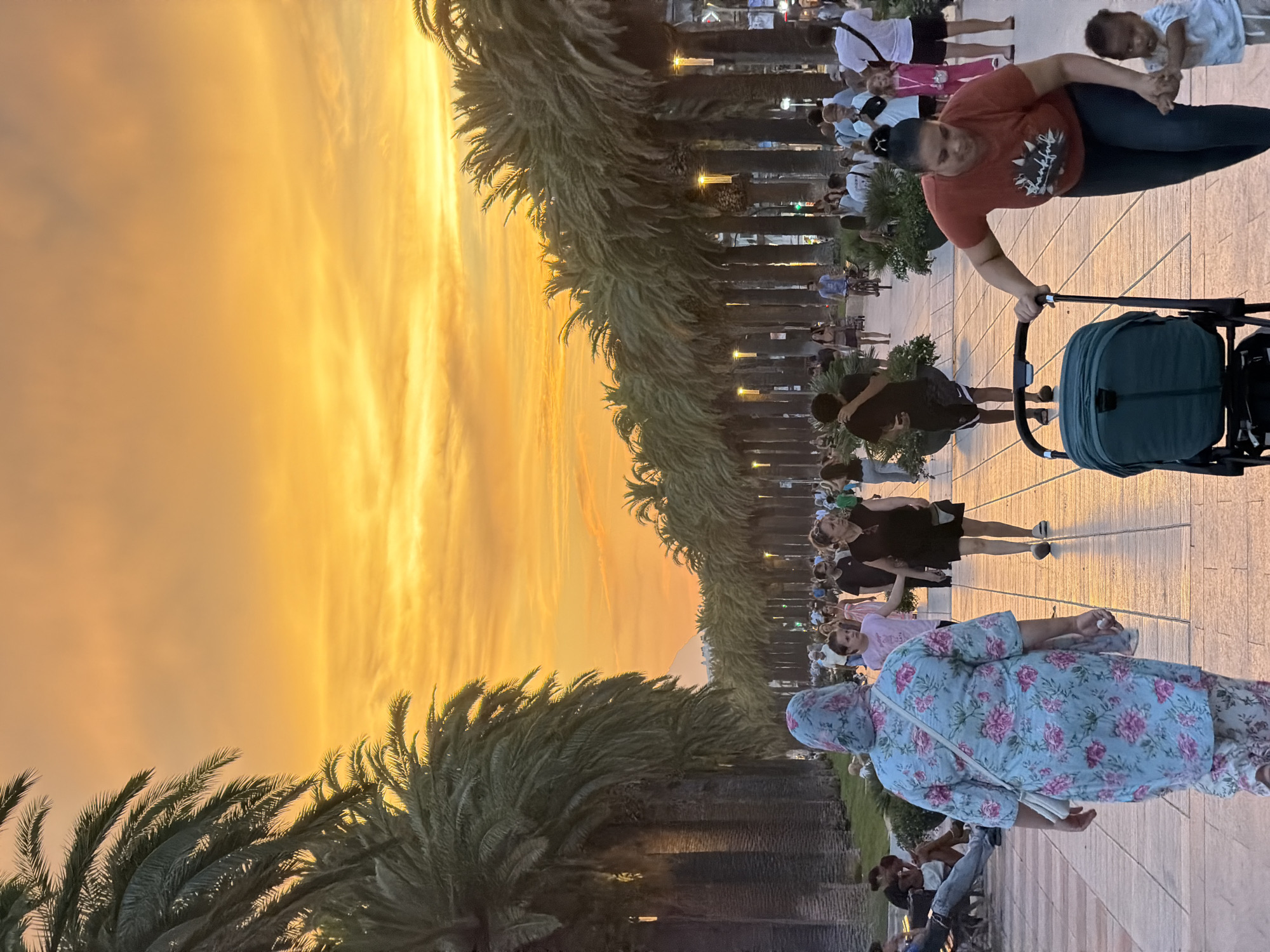 This image depicts a wide pedestrian promenade lined with tall palm trees, likely located in the city of Salou, Spain, given the context. The sky above is a vibrant orange and yellow, suggesting a sunset or sunrise. Several people are walking along the promenade, some individually and others in groups. In the foreground, a woman is pushing a dark teal stroller, with a young child holding her hand beside her. Further down the promenade, other individuals and groups are visible, with some seated on benches. The palm trees cast long shadows on the paved walkway, and decorative lights are visible along the avenue. The overall atmosphere appears to be one of leisure and outdoor activity.