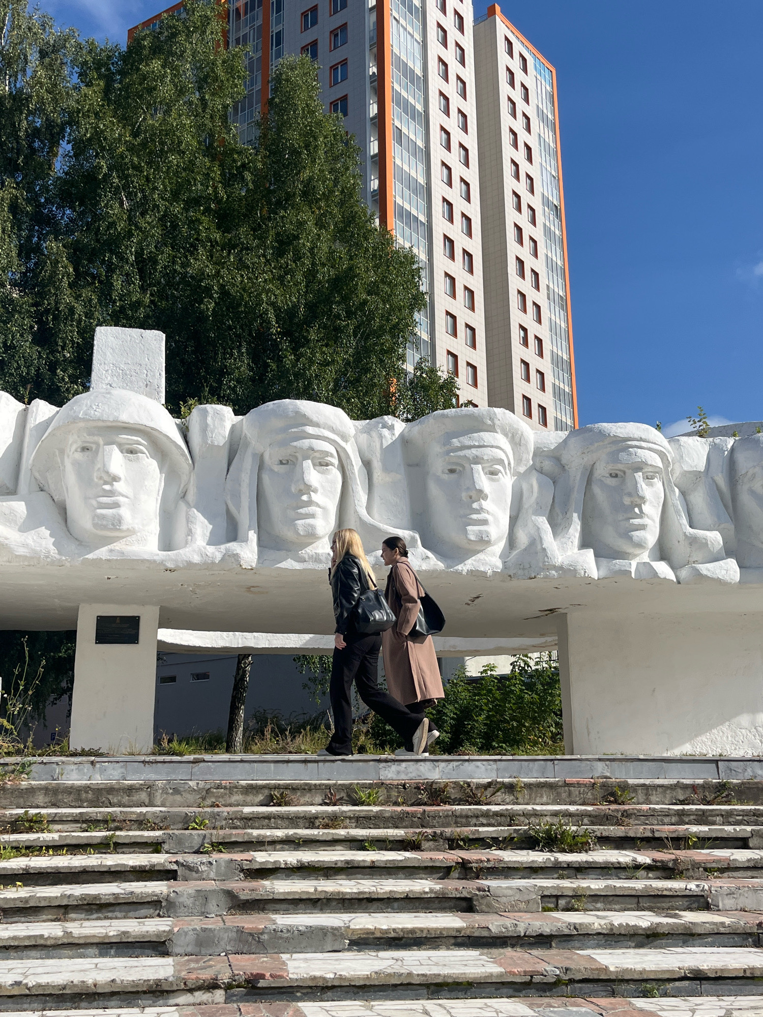 The image shows a monumental sculpture depicting several large, white, stylized human heads. These heads appear to be made of concrete or plaster and are part of an outdoor monument. Two women are walking on a set of worn, stone stairs in front of the sculpture. In the background, a tall, modern apartment building with orange and white detailing stands against a clear blue sky, and lush green trees are visible. The sculpture is located in the city of Perm, Russia, specifically in front of the memorial complex "Perm 1 - Perm 2" on Komsomolsky Prospekt. One of the women is wearing a black leather jacket and black pants, while the other is wearing a beige coat and black pants. Both are carrying bags. The monument itself features at least four prominent heads, one wearing a military-style helmet, and others with head coverings resembling a keffiyeh. The stairs are weathered and have small plants growing between the stone slabs.