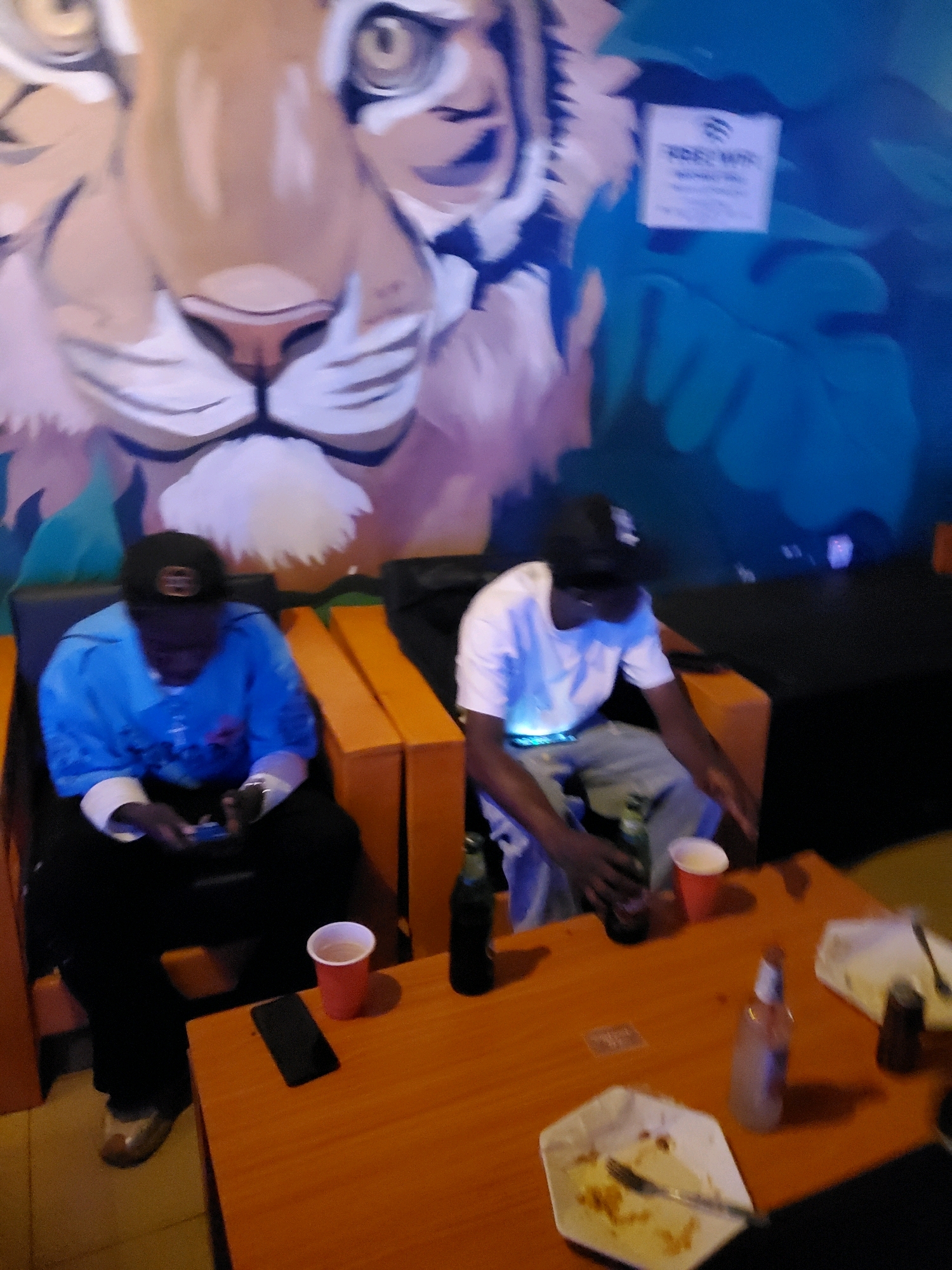 The image depicts two individuals seated on orange cushioned benches in what appears to be an indoor establishment. A large mural of a lion's face is visible on the wall behind them. One individual, on the left, wears a blue long-sleeved shirt and black pants, holding a phone. The other, on the right, is in a white t-shirt and light-colored pants, holding a beer bottle. A wooden table sits in front of them, with various items including a red cup, a phone, beer bottles, and used paper plates. The establishment is likely a bar or lounge, possibly located in Jos, Nigeria, given the context provided. The lighting suggests an evening or dimly lit environment.